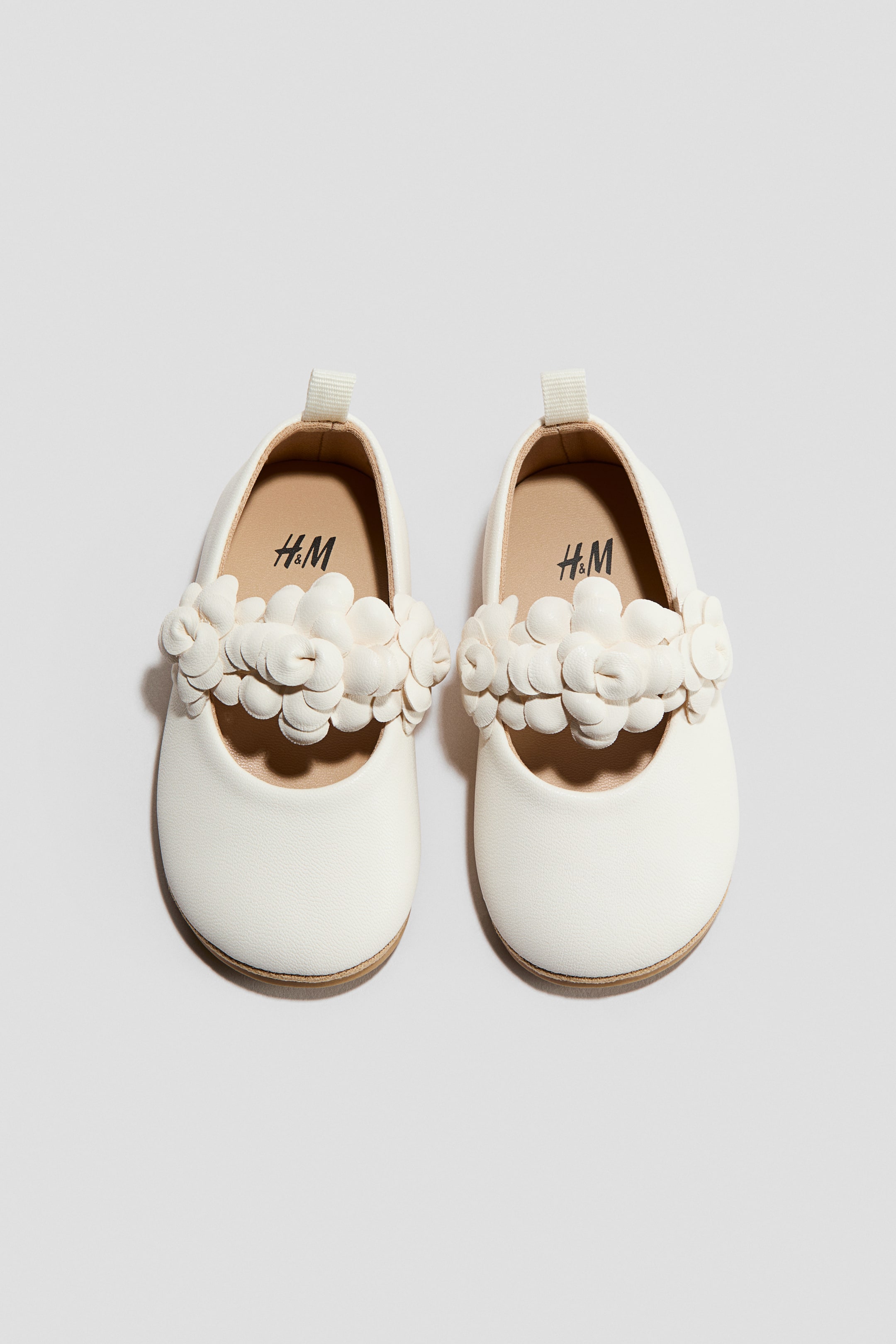 View larger image: A pair of off-white baby ballet flats is facing FRONT, displaying multiple 3D floral embellishments across the instep of each shoe. The shoes are also featuring small fabric pull tabs at the back and light brown soles.