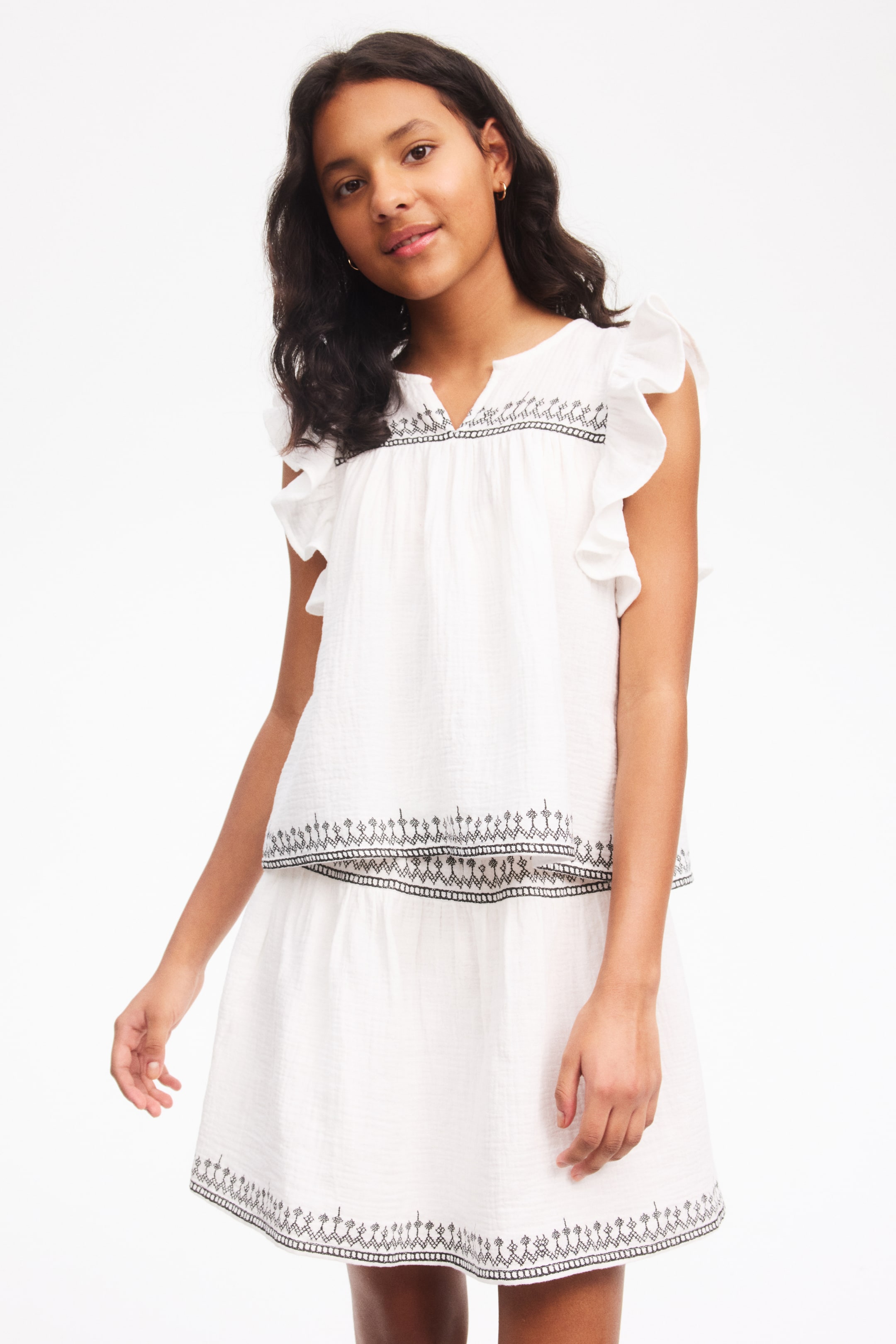 View larger image: A white crinkled cotton top and matching skirt set faces forward, featuring a notched V-neckline, ruffle cap sleeves and black embroidered trim along the top's yoke and hem. The tiered skirt showcases matching black embroidery above its hem.