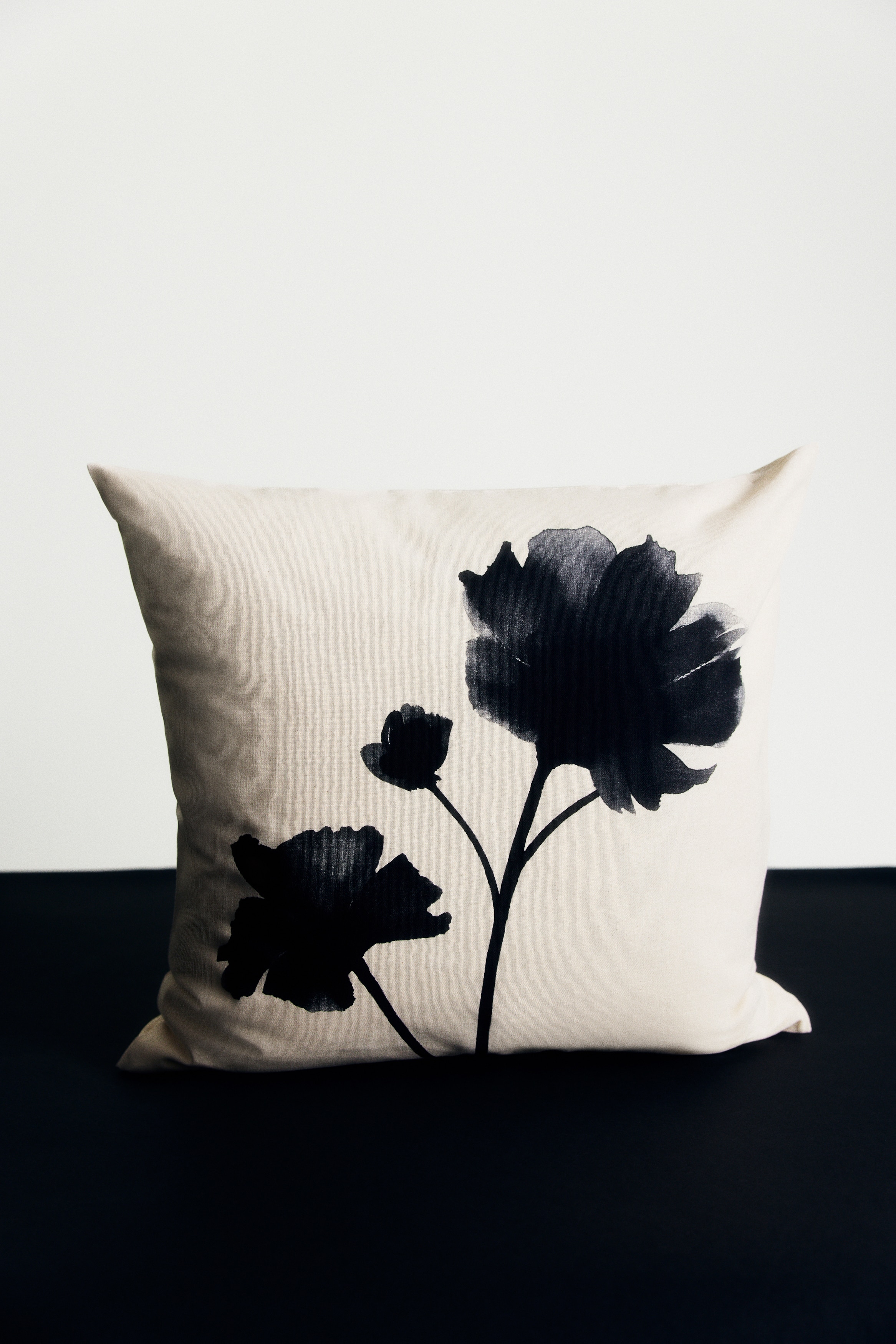 A square scatter cushion features a black floral print on a light beige fabric, positioned at a slight angle to reveal its depth. Three black flowers, depicted in an ink wash style with visible stems, are arranged across the front surface.