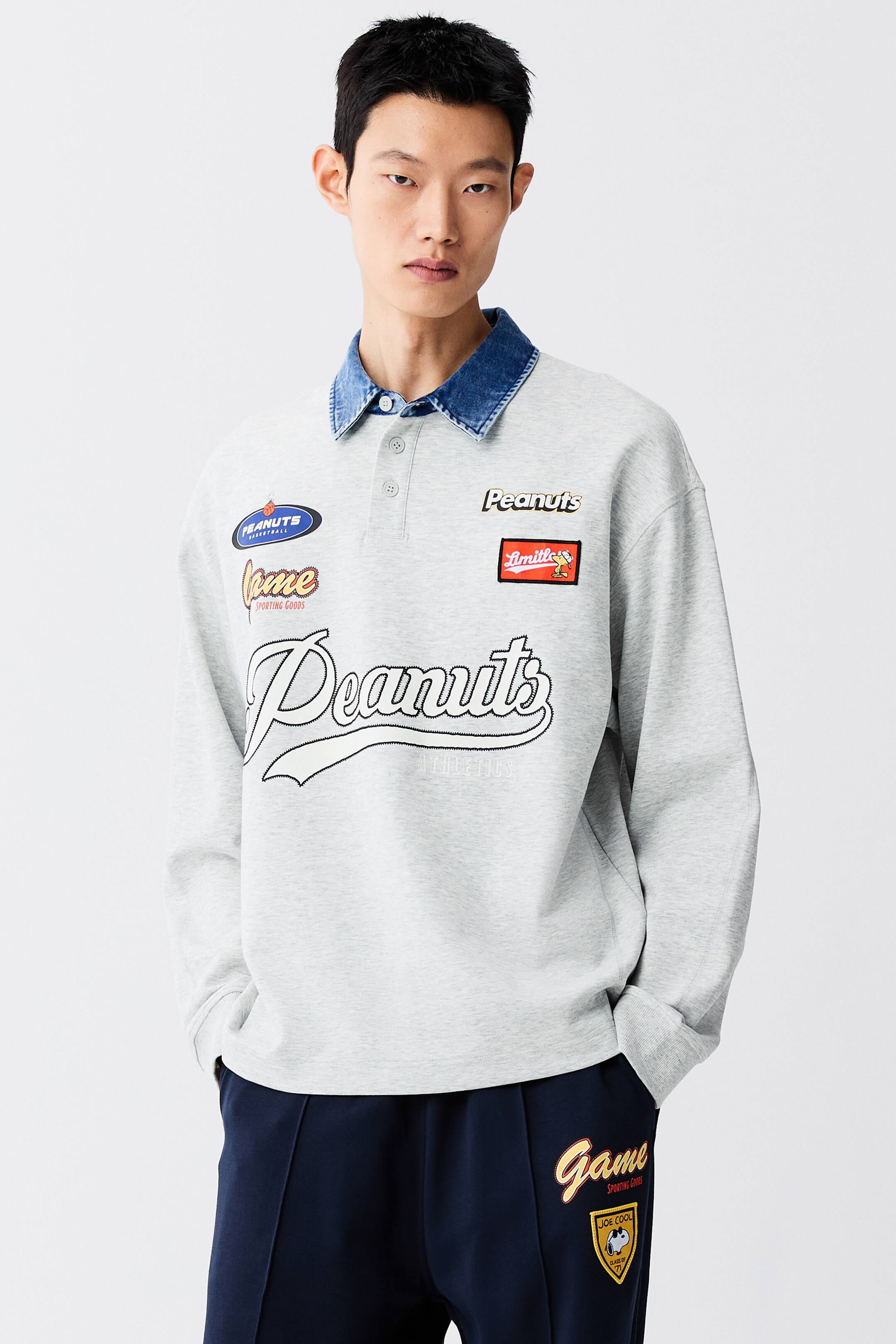 View larger image: A light grey rugby shirt featuring various graphic prints and patches is being worn by the model over a blue denim collared shirt. The loose-fit, long-sleeved shirt is styled with dark blue sweatpants featuring coordinating graphics.