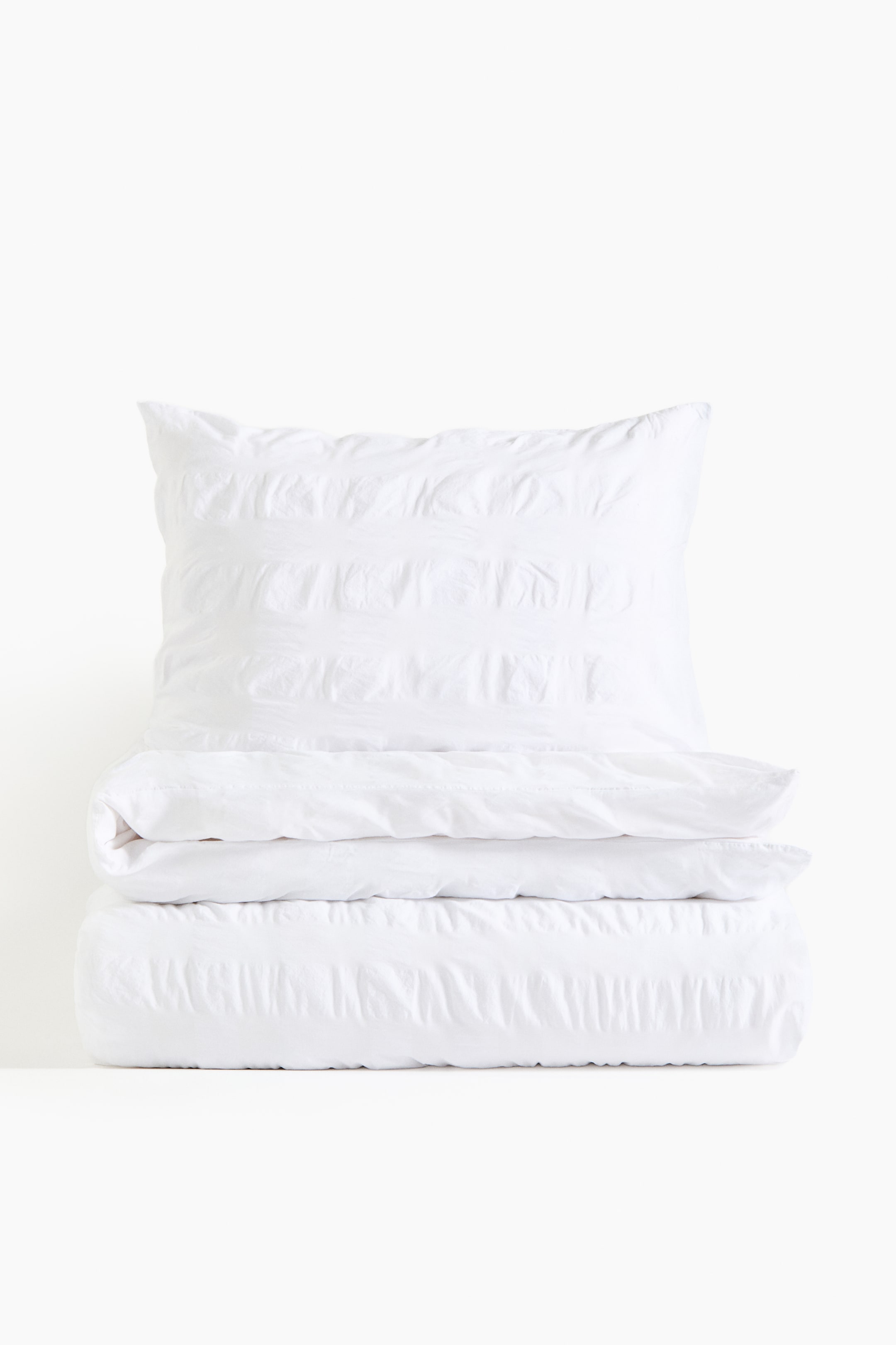 Ver imagem maior: Um conjunto de roupa de cama branca está apresentado em pilha, com uma almofada colocada na vertical no topo. Uma capa de edredão dobrada está posicionada abaixo da almofada, e outra capa de edredão dobrada encontra-se na parte inferior, todas exibindo faixas horizontais com textura enrugada.