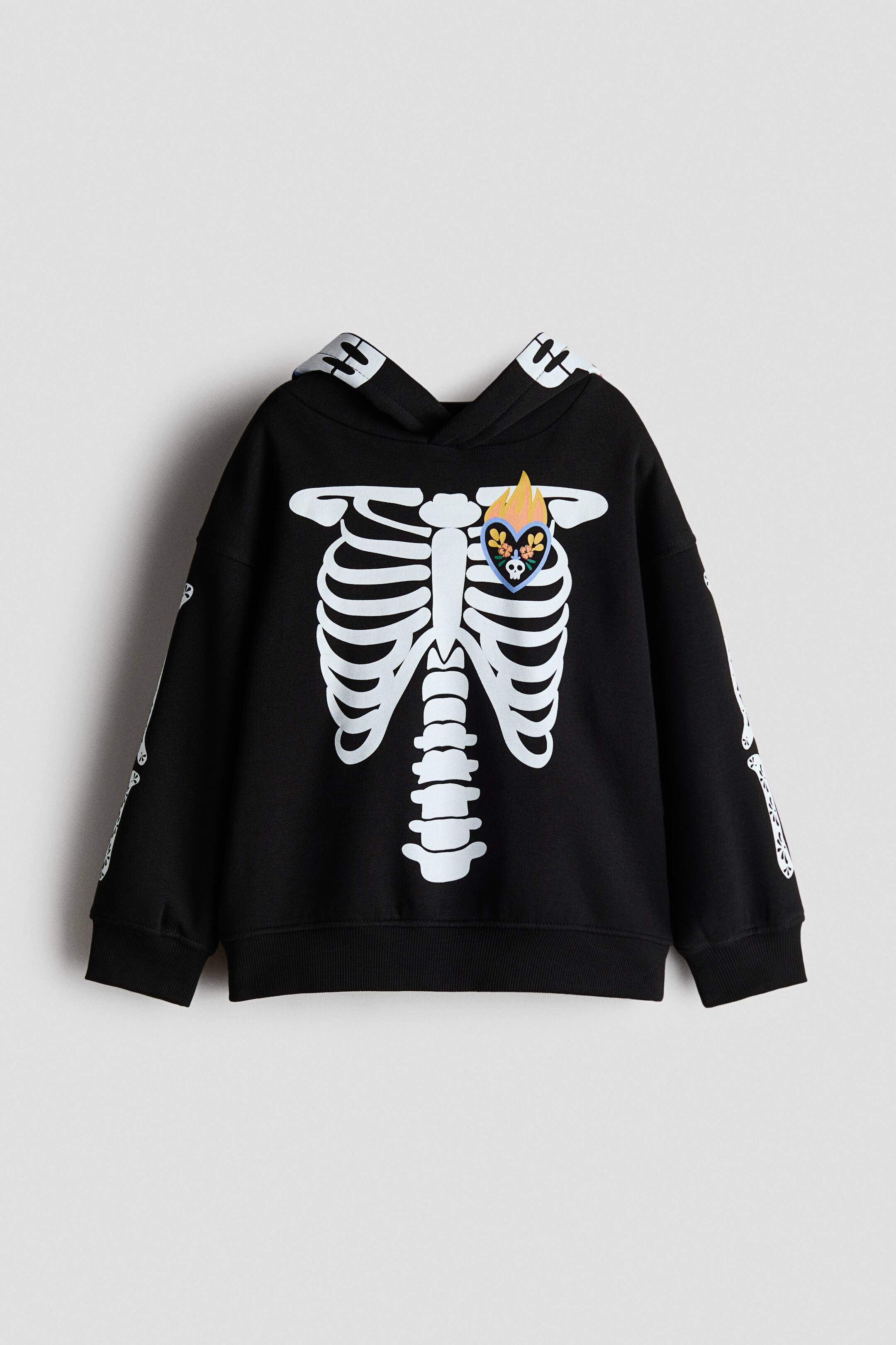View larger image: A black hoodie features a white skeleton rib cage and spine print on the front, with a colorful heart patch on the left chest depicting a skull and flowers, topped with yellow and orange flames. White skeleton arm bones are printed along the sleeves, and the hoodie includes ribbed cuffs, a ribbed hem, and a hood with a white patterned interior lining.