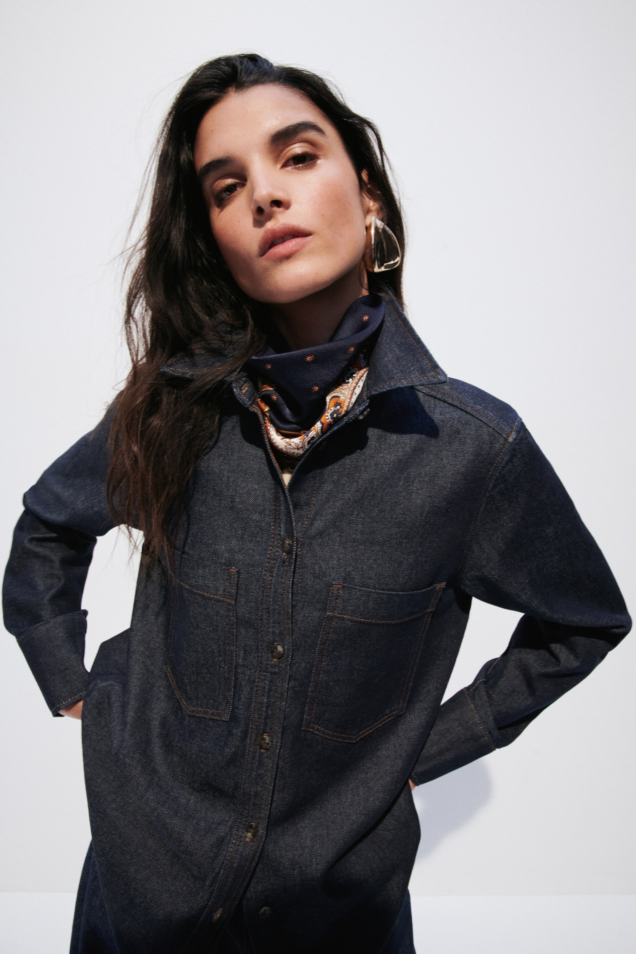 View larger image: The dark wash denim shirtdress features a pointed collar, a full button-front closure with brown buttons, and contrasting light brown stitching outlining its two flapped patch pockets on the chest and two patch pockets on the lower front. Its long sleeves are rolled up at the cuffs, and a patterned scarf is visible at the neckline.