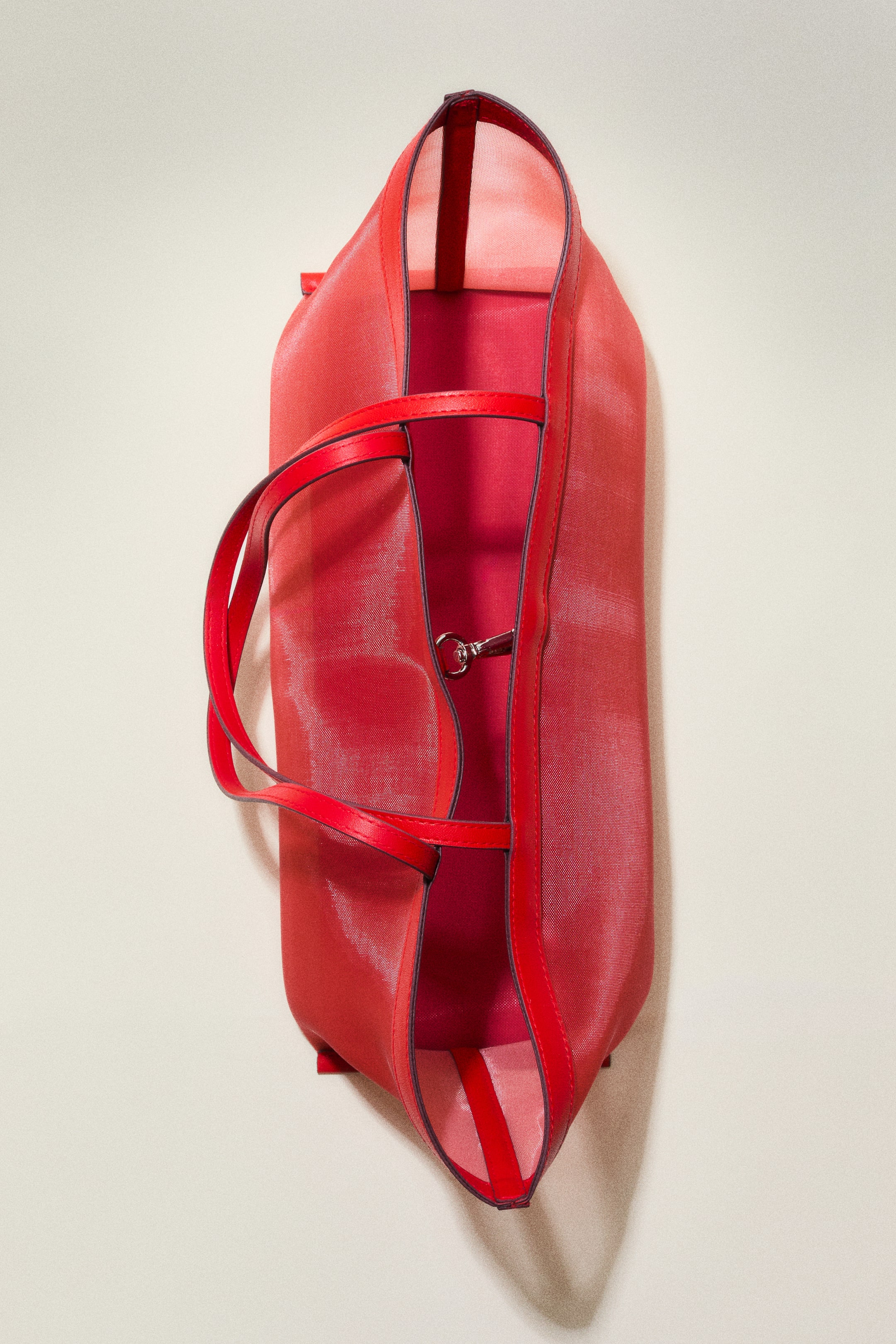 Agrandir l'image: Un sac cabas rouge est présenté en vue plongeante, légèrement inclinée, et se tient debout. Le sac présente un corps en maille avec des détails en tissu enduit soulignant les bords et formant les deux poignées supérieures. Une fermeture à crochet en métal relie l'ouverture supérieure du sac.
