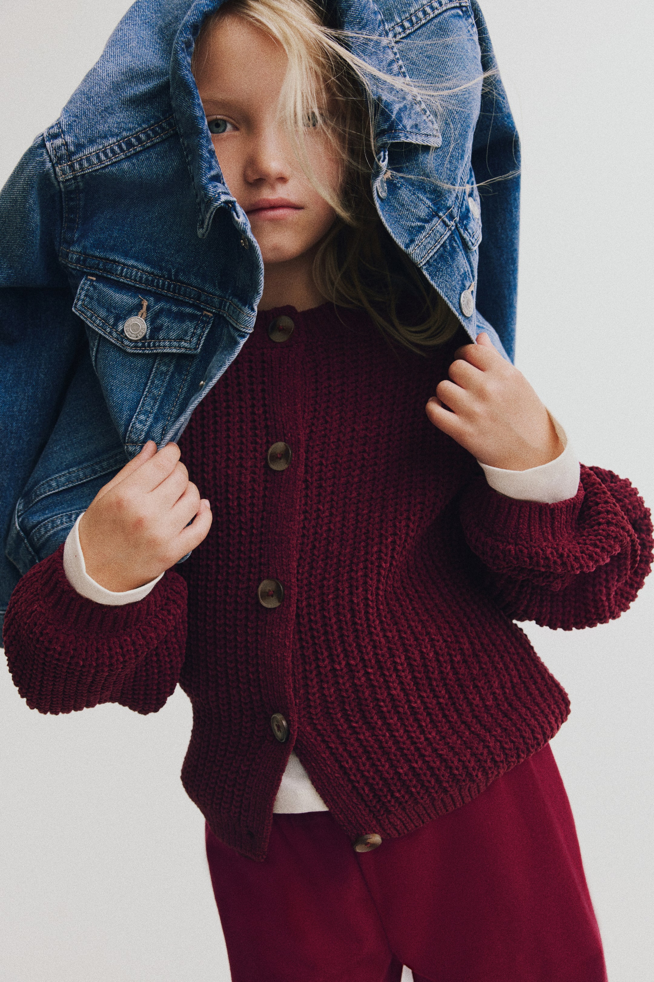 Ver imagem maior: Estas calças de fato de treino cor borgonha, mostradas de frente, apresentam um corte descontraído de perna larga em tecido sweatshirt suave. Estão combinadas com um cardigan de malha com botões em borgonha a condizer e uma jacket de ganga azul colocada sobre a cabeça da modelo.