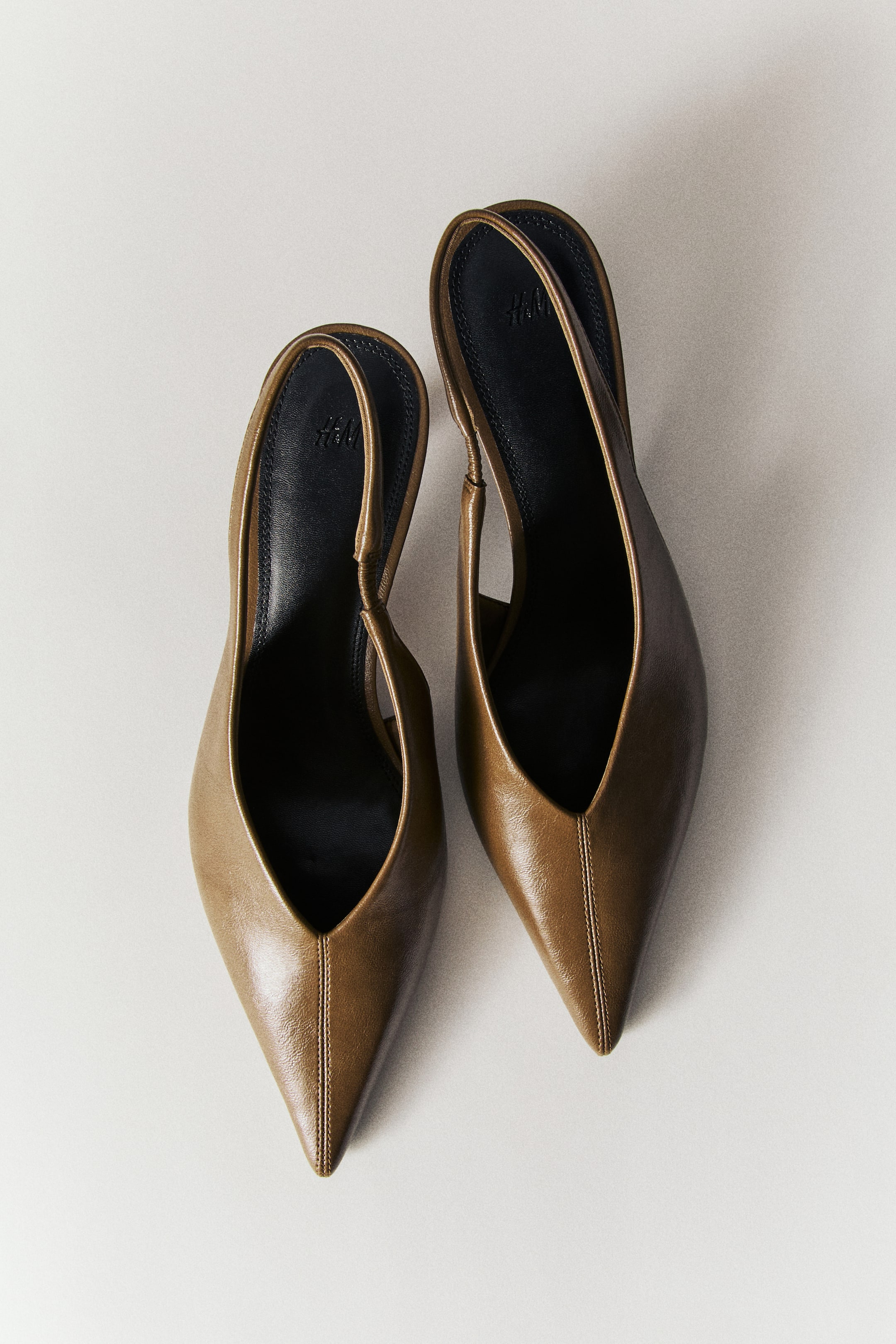 View larger image: Slingback shoes in a smooth olive-brown material are shown from a top-down view, featuring sharp pointed toes with a central vertical seam. The design includes narrow heel straps with small elastic inserts and contrasting black insoles, with the low kitten heels partially visible underneath.