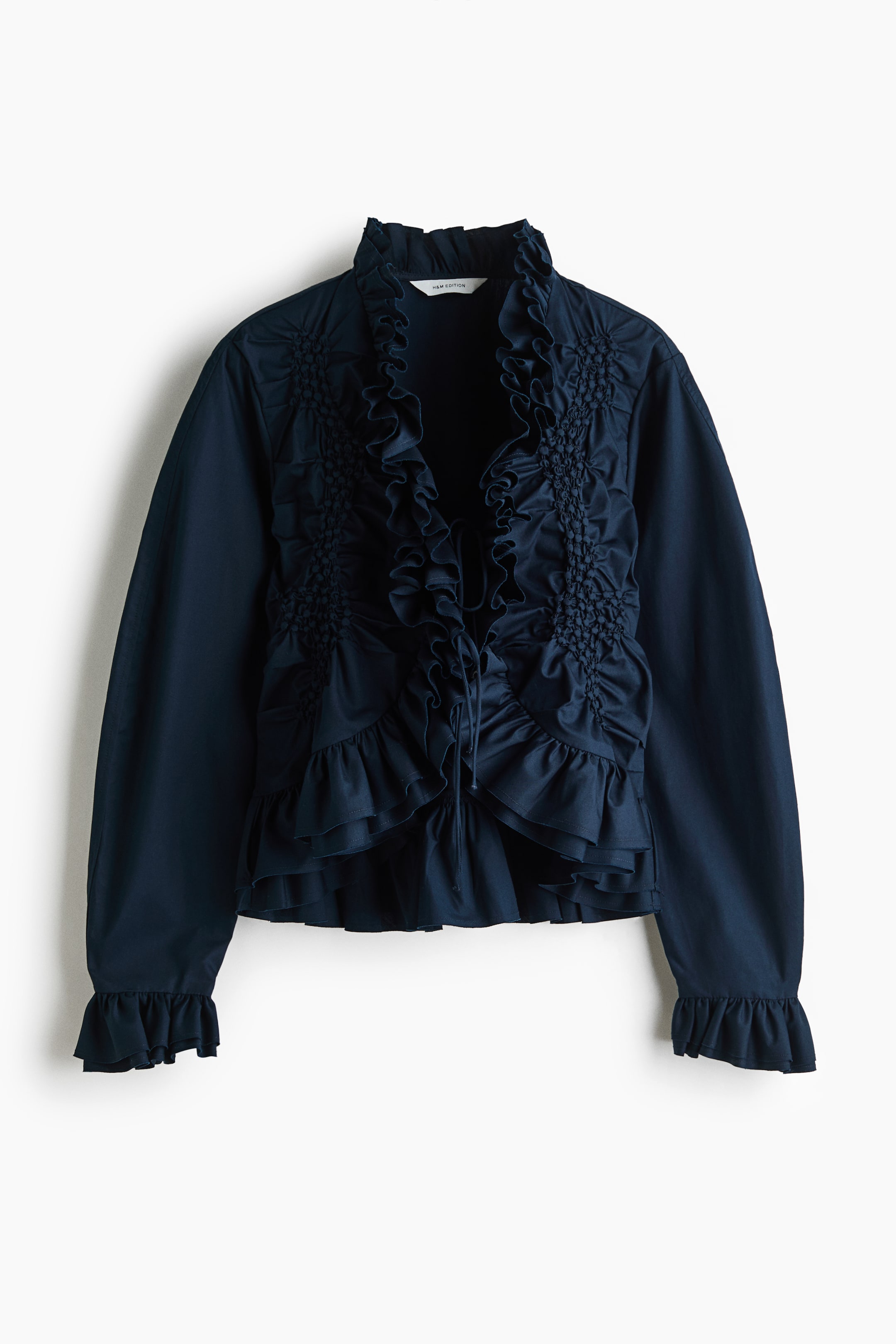 View larger image: A dark blue blouse is shown from the front, crafted from a crisp cotton weave and featuring a deep V-neckline with an open front secured by two sets of narrow ties. The front is detailed with smocking, while a double-layered frill trim adorns the neckline, continues down the front, encircles the hem, and finishes the cuffs of the long, voluminous sleeves.