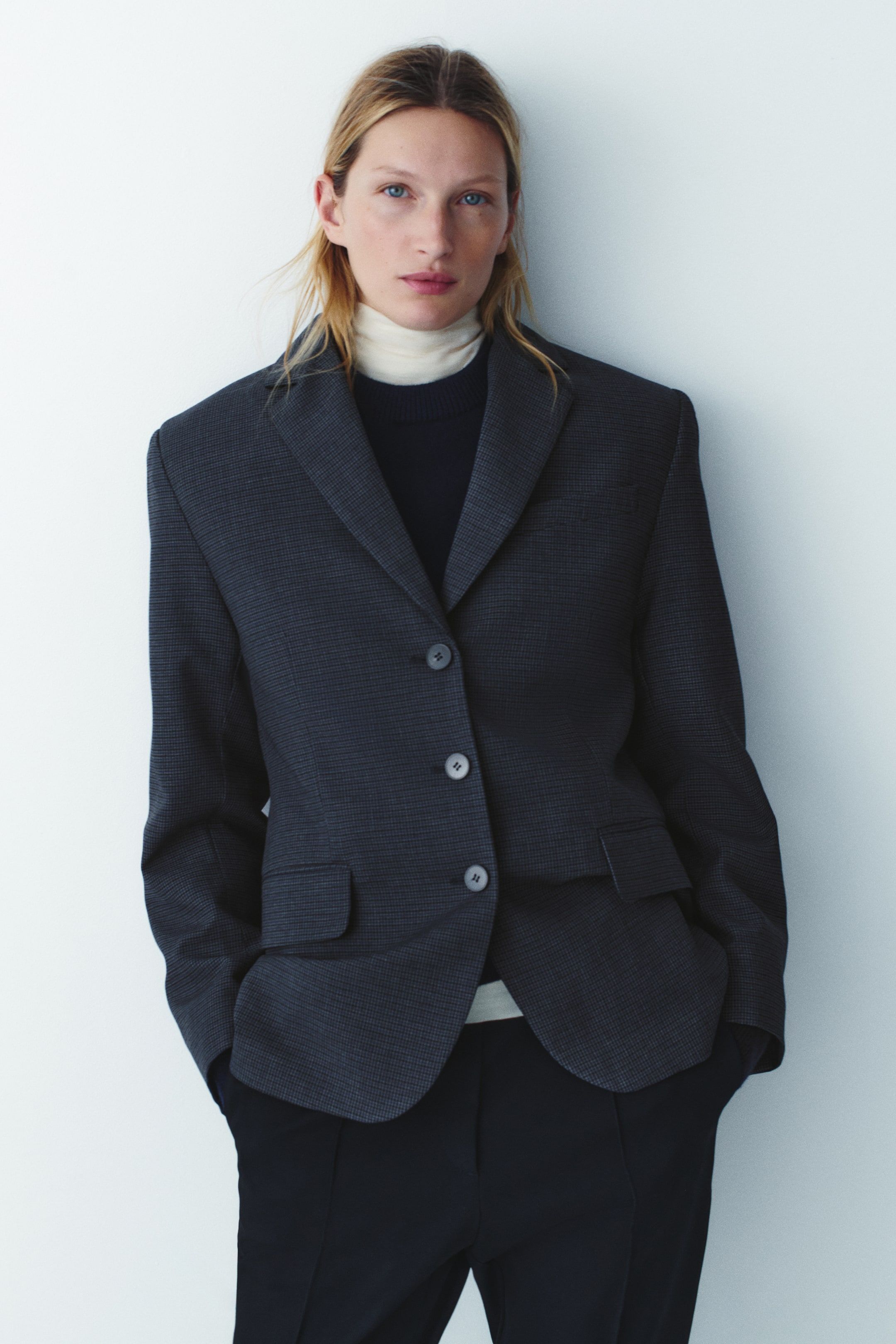 View larger image: The model is wearing a dark gray patterned twill single-breasted blazer layered over a black sweater, a cream turtleneck, and dark trousers. The blazer features structured shoulders, notch lapels, a three-button front closure, and front flap pockets.