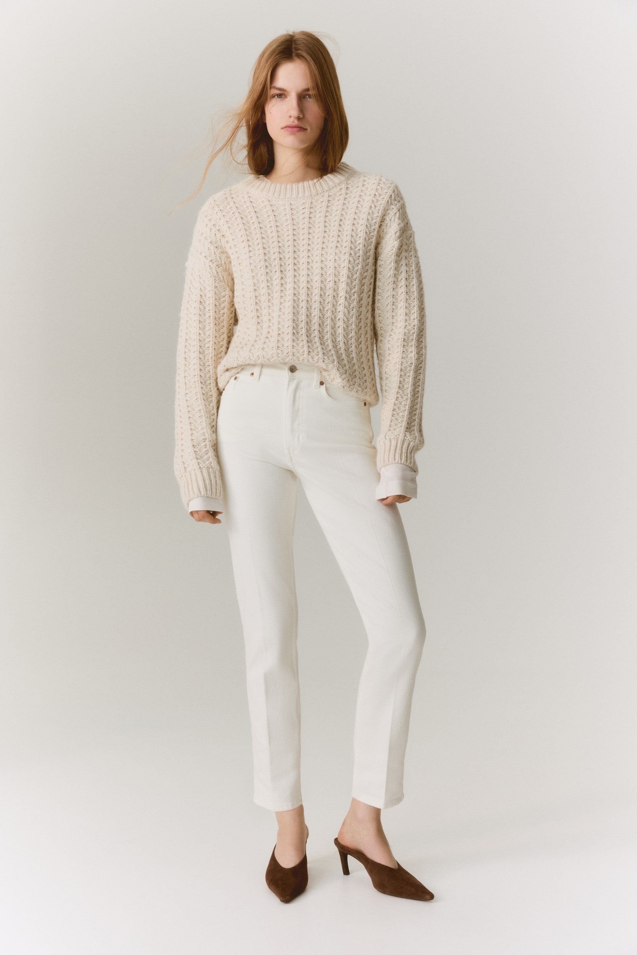 Zobraziť väčší obrázok: Krémový pletený sveter je prezentovaný s výhľadom spredu, vyznačujúci sa hrubým, textúrovaným pleteným vzorom a okrúhlym výstrihom. Jeho dlhé, voľné rukávy končia pri zápästiach, s bielymi manžetami košele viditeľne navrstvenými pod nimi.