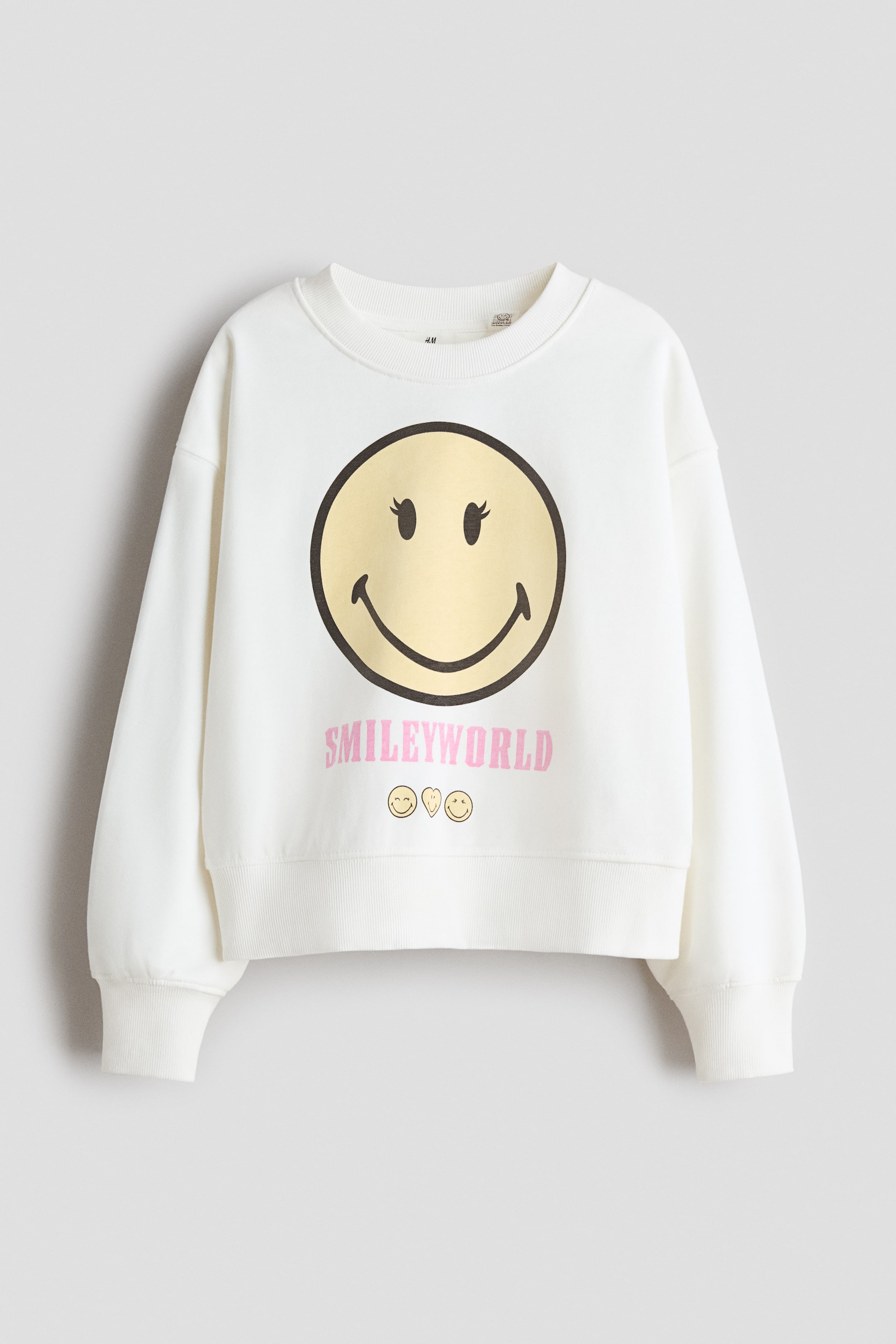View larger image: An oversized cream sweatshirt is featuring a large yellow smiley face print with black outlines and small pink eyelashes on the FRONT. 