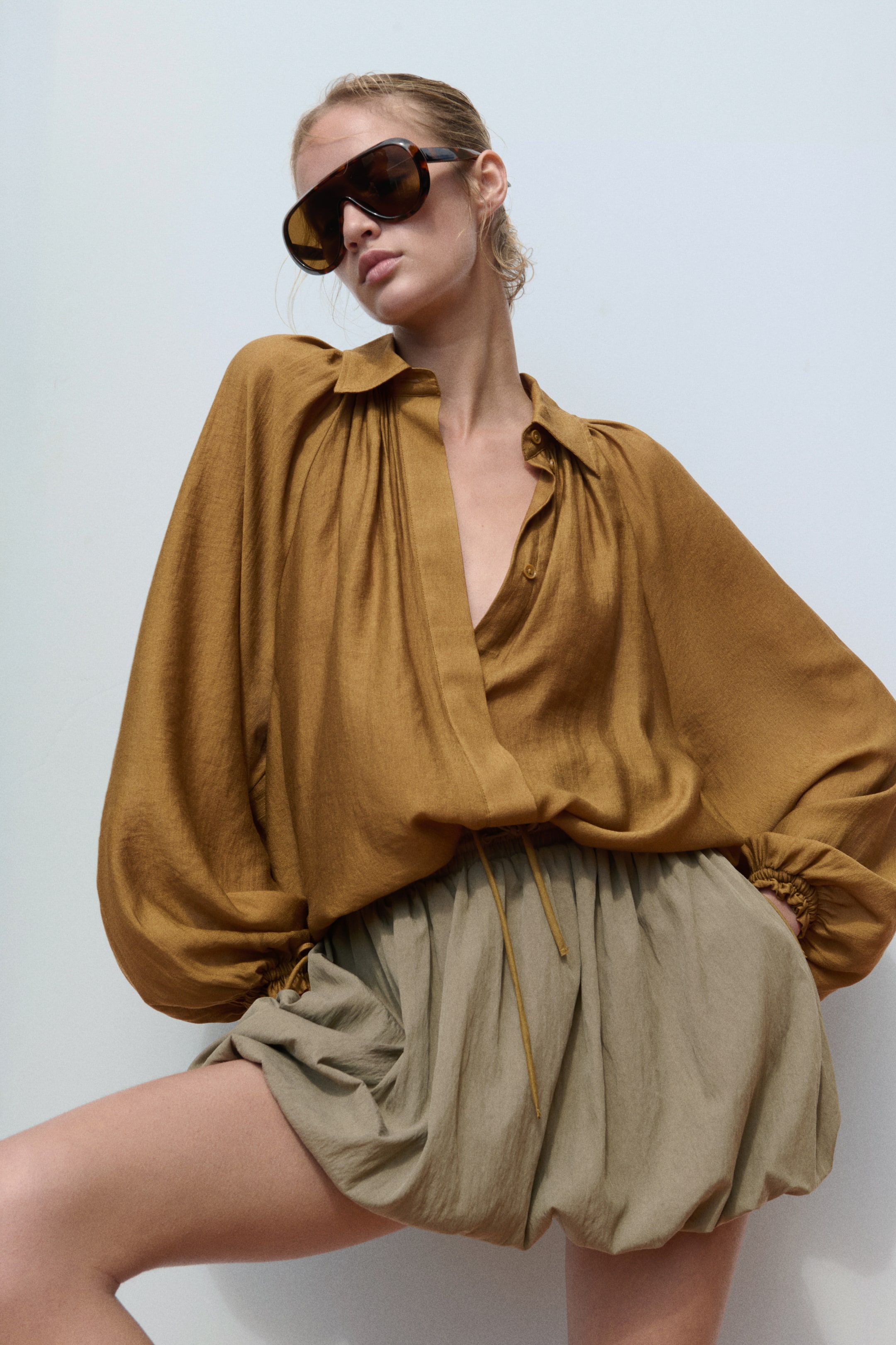 View larger image: The model is wearing an oversized shirt in a golden-brown viscose blend. It showcases a collar, gathers at the neckline, and long raglan sleeves with drawstrings at the cuffs and hem, creating a voluminous silhouette over olive green shorts.