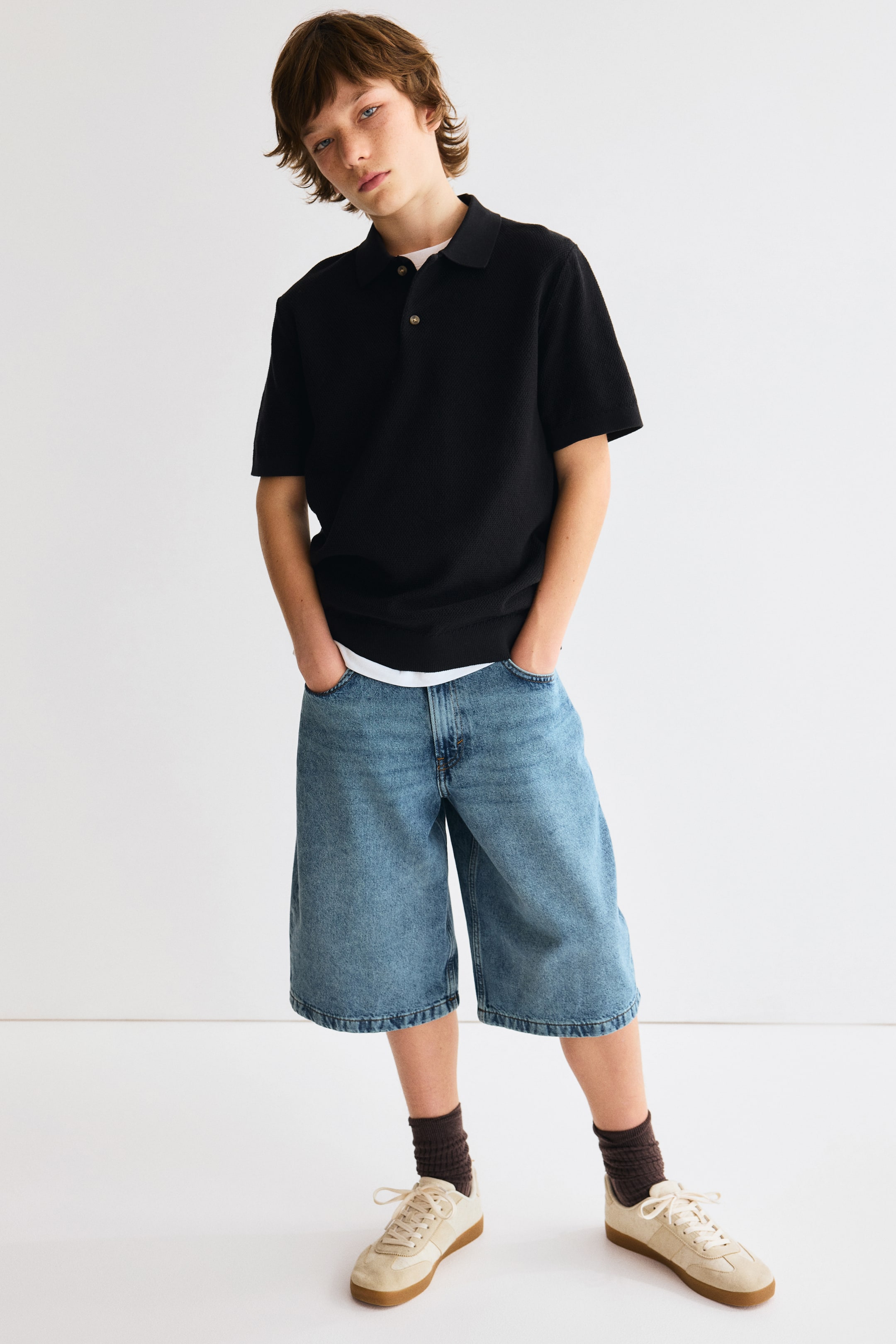Agrandir l'image: Un polo noir à manches courtes présente un tissu en maille texturée, un col polo et une patte de boutonnage à deux boutons avec des boutons marron clair. La chemise à coupe décontractée est superposée sur un t-shirt blanc, avec son ourlet blanc visible.