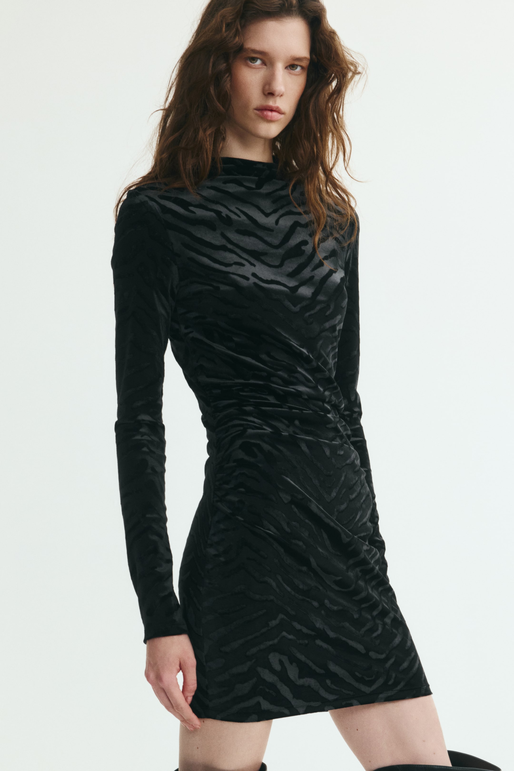 Vezi imaginea mai mare: O rochie mini neagră, mulată, cu un model zebra texturat, este purtată de model. Rochia este concepută cu guler tunică, mâneci lungi și detalii încrețite de-a lungul cusăturilor laterale, peste șolduri.
