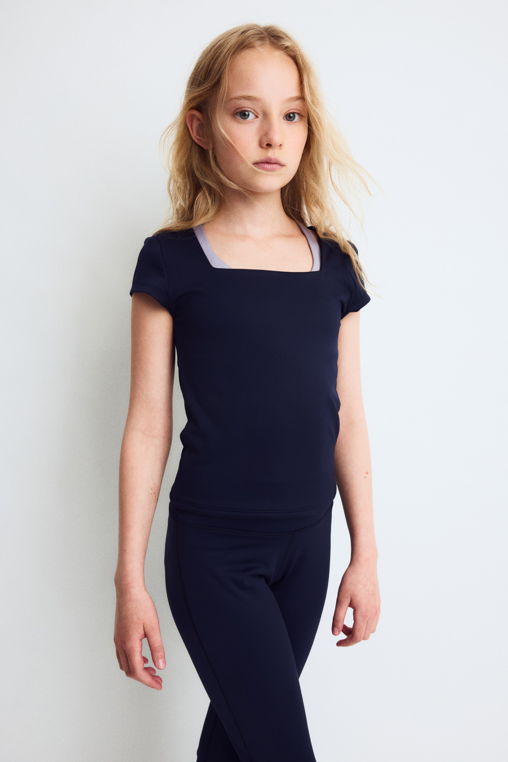 Agrandir l'image: Un ensemble de sport bleu marine est présenté de face, composé d’un haut moulant à manches courtes avec un décolleté carré et d’un legging assorti. Des bretelles lilas clair provenant d’un sous-vêtement superposé sont visibles au niveau de l’encolure.