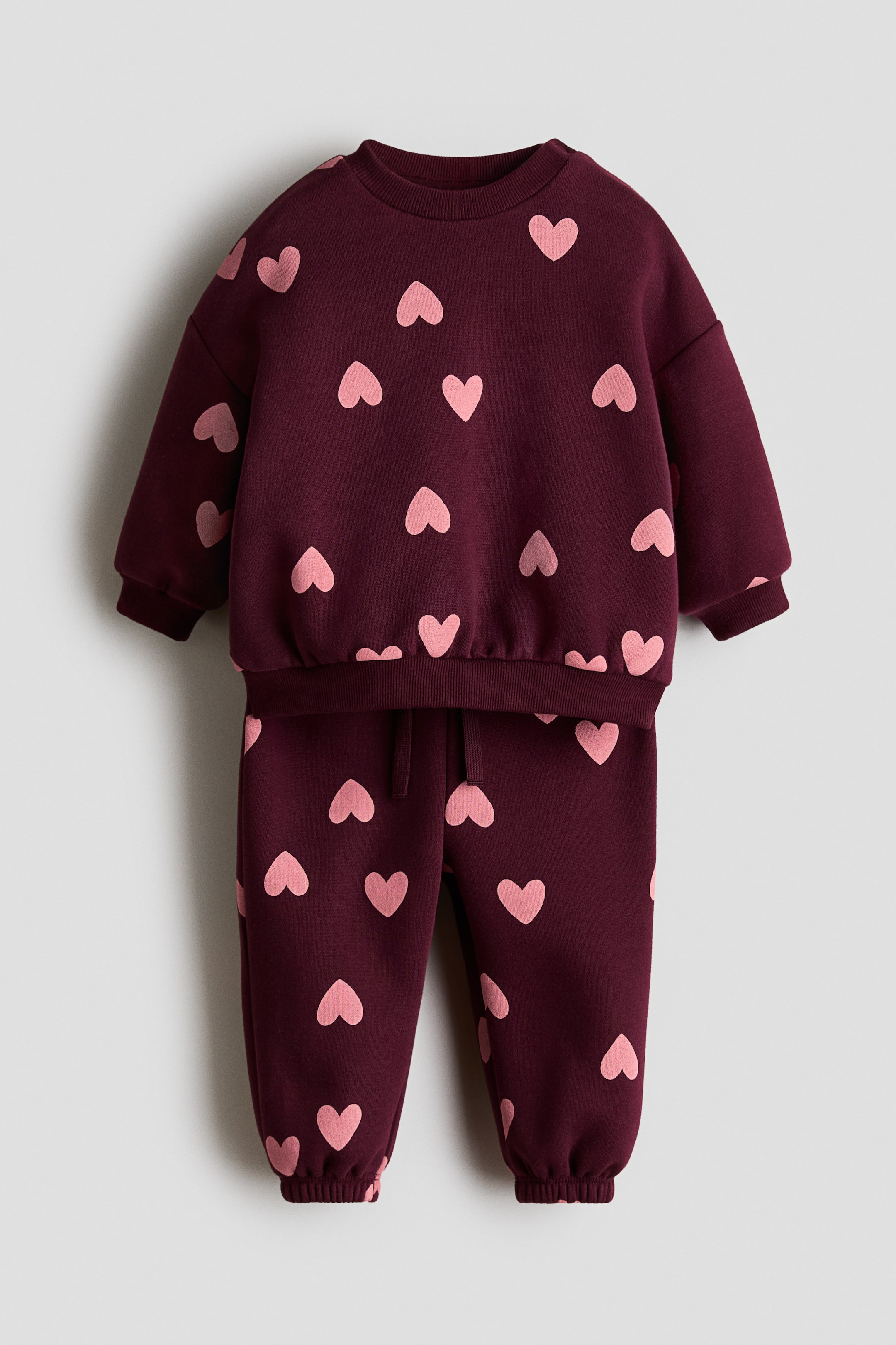 View larger image: A burgundy two-piece set is being showcased, consisting of a sweatshirt and sweatpants, both adorned with a scattered pink heart pattern. The sweatshirt features a crew neck, long sleeves with ribbed cuffs, and a ribbed hem, while the sweatpants boast an elasticated drawstring waistband with a visible drawstring and ribbed ankle cuffs.