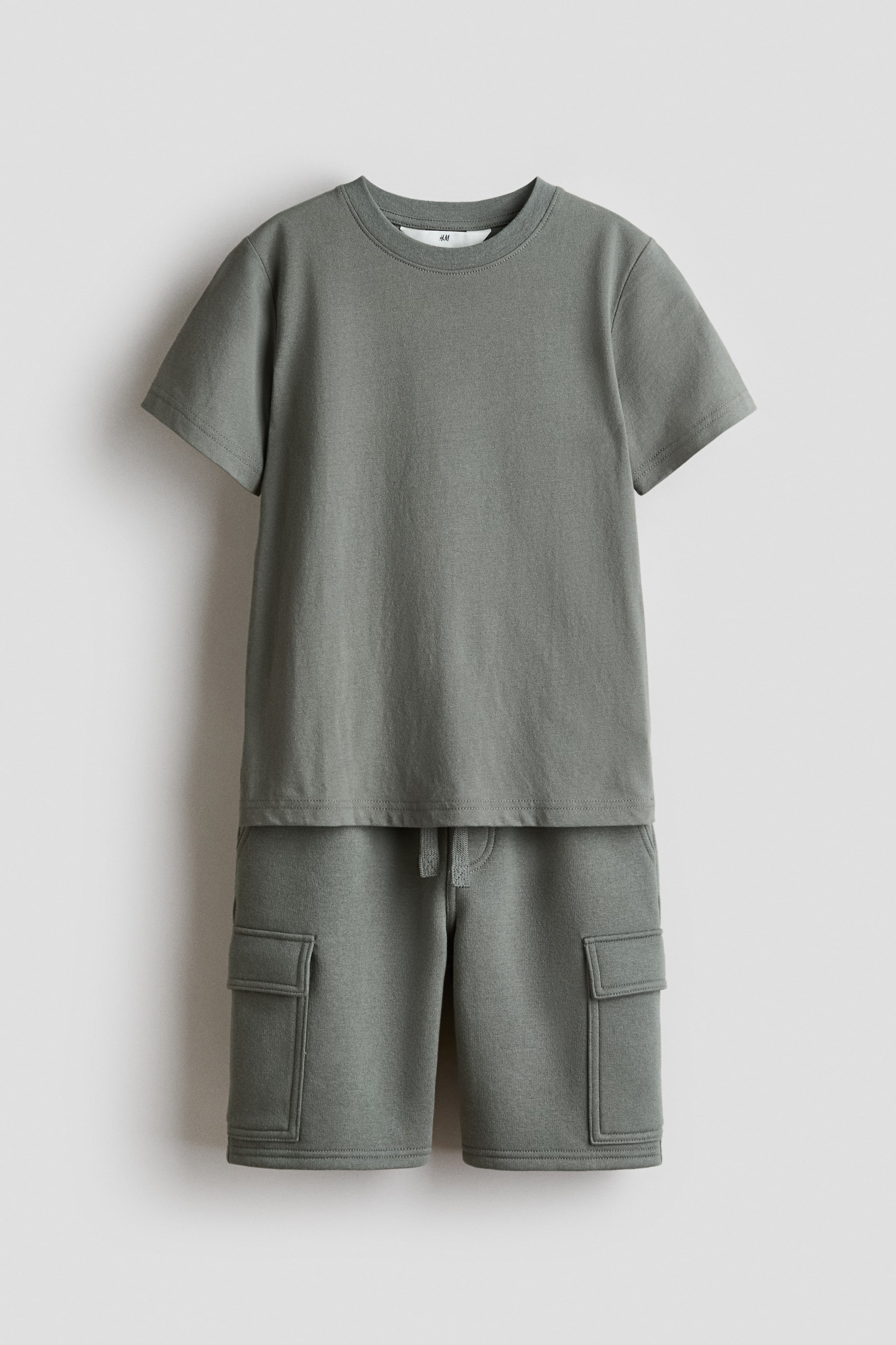 View larger image: A front-facing set showcases a short-sleeved, muted green T-shirt with a round, rib-trimmed neckline and a straight-cut hem. The matching shorts feature an elasticated waist with a visible drawstring and patch leg pockets with flaps on both legs.