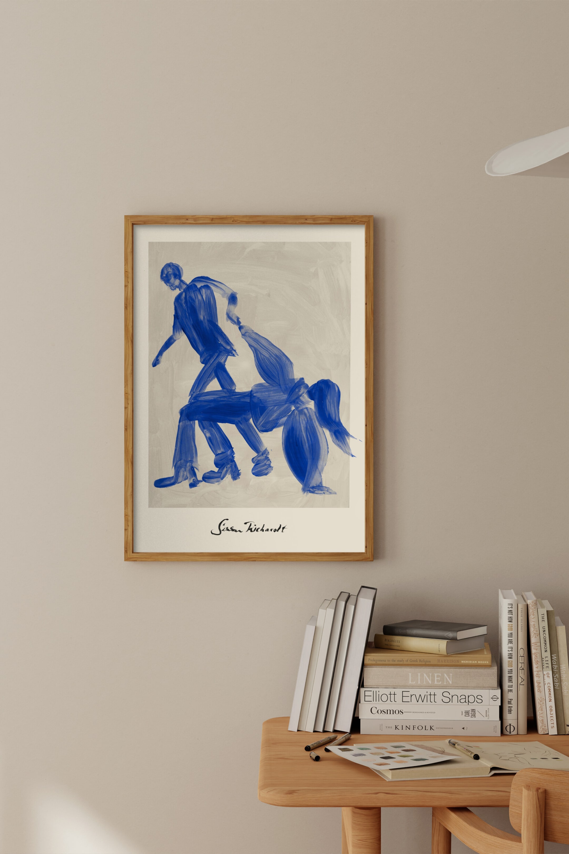 Se større billede: Et indrammet kunsttryk hænger lodret, med en lys træramme og hvidt passepartout. Trykket viser en illustration af to figurer, gengivet i ekspressive blå penselstrøg mod en lys grå baggrund, hvor den ene figur står og ser ud til at trække en anden figur i en sammenbøjet position, og kunstnerens signatur "Sissan Kicharolt" er synlig nedenfor.