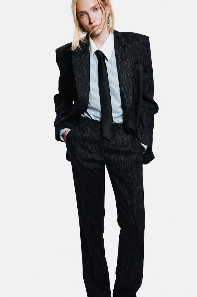 A dark charcoal pinstriped suit is being presented, featuring an oversized blazer with broad, structured shoulders and notch lapels, worn unbuttoned over a light blue striped shirt and a black tie, and matching straight-leg trousers. - 1
