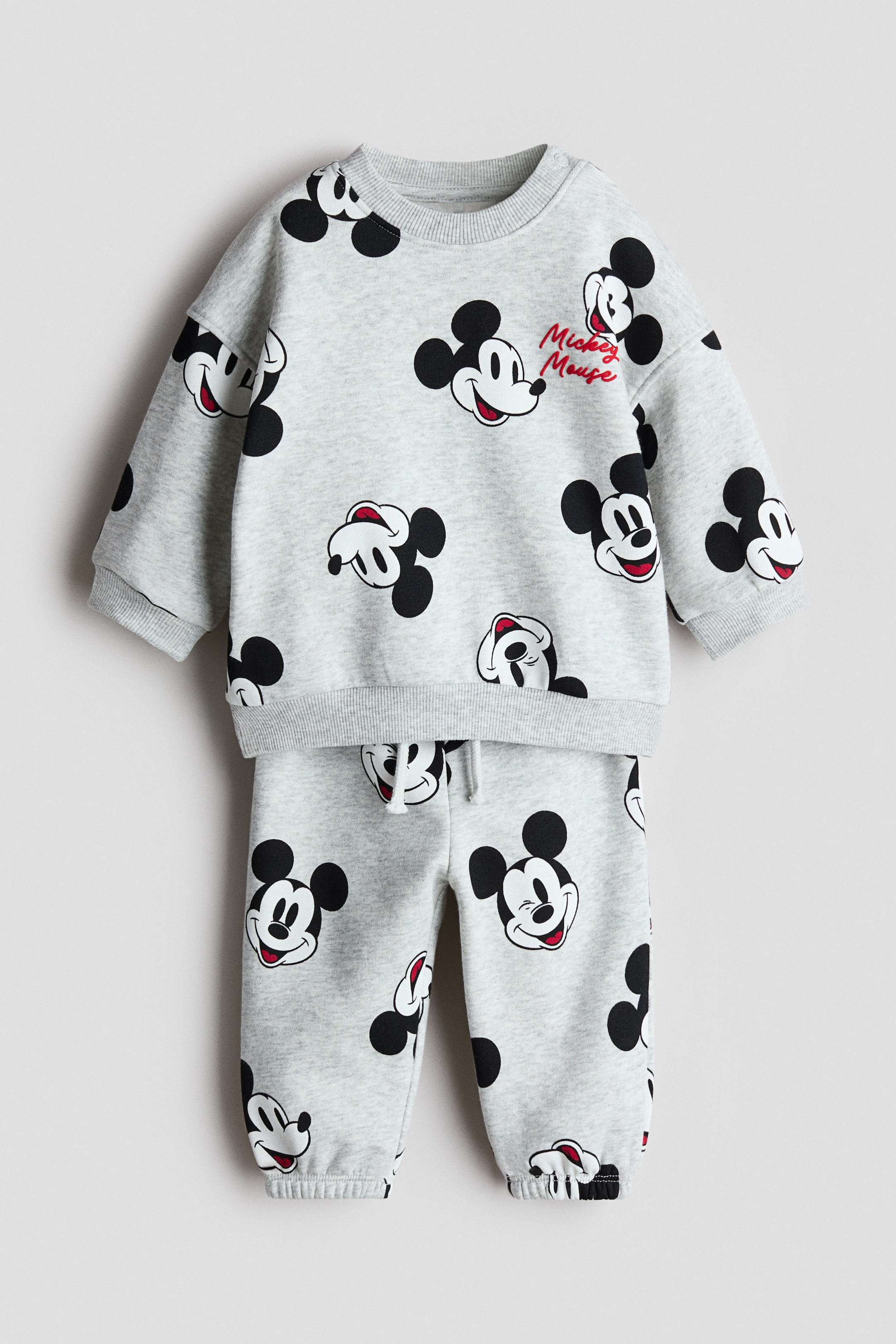 Ver imagen más grande: Un conjunto de sudadera estampada de 2 piezas se presenta de frente, con una tela de marl gris claro y un estampado completo de caras de Mickey Mouse en blanco y negro. La sudadera con cuello redondo muestra puños acanalados y un dobladillo acanalado, con pequeños botones a presión visibles en el hombro izquierdo y el texto rojo 'Mickey Mouse' en el pecho. Los pantalones de chándal a juego completan el conjunto, mostrando una cintura elástica con cordón y puños acanalados en los tobillos.