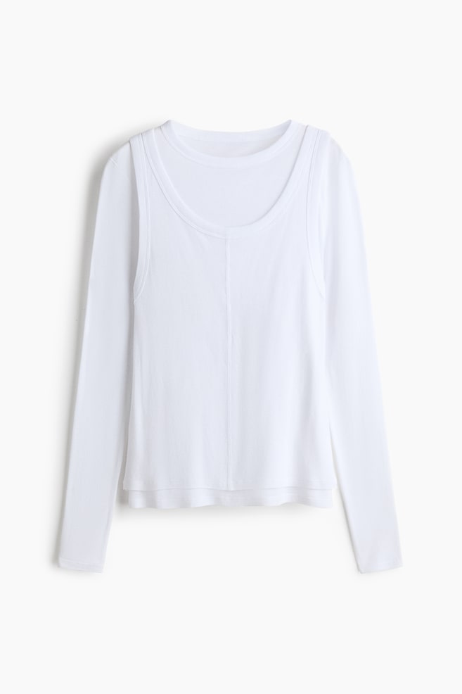 A white layered top is shown from the front, featuring a long-sleeve crew-neck base layer beneath a scoop-neck tank top. The outer tank top features a vertical seam down the centre front and a slightly shorter hemline, creating a tiered effect. - 1