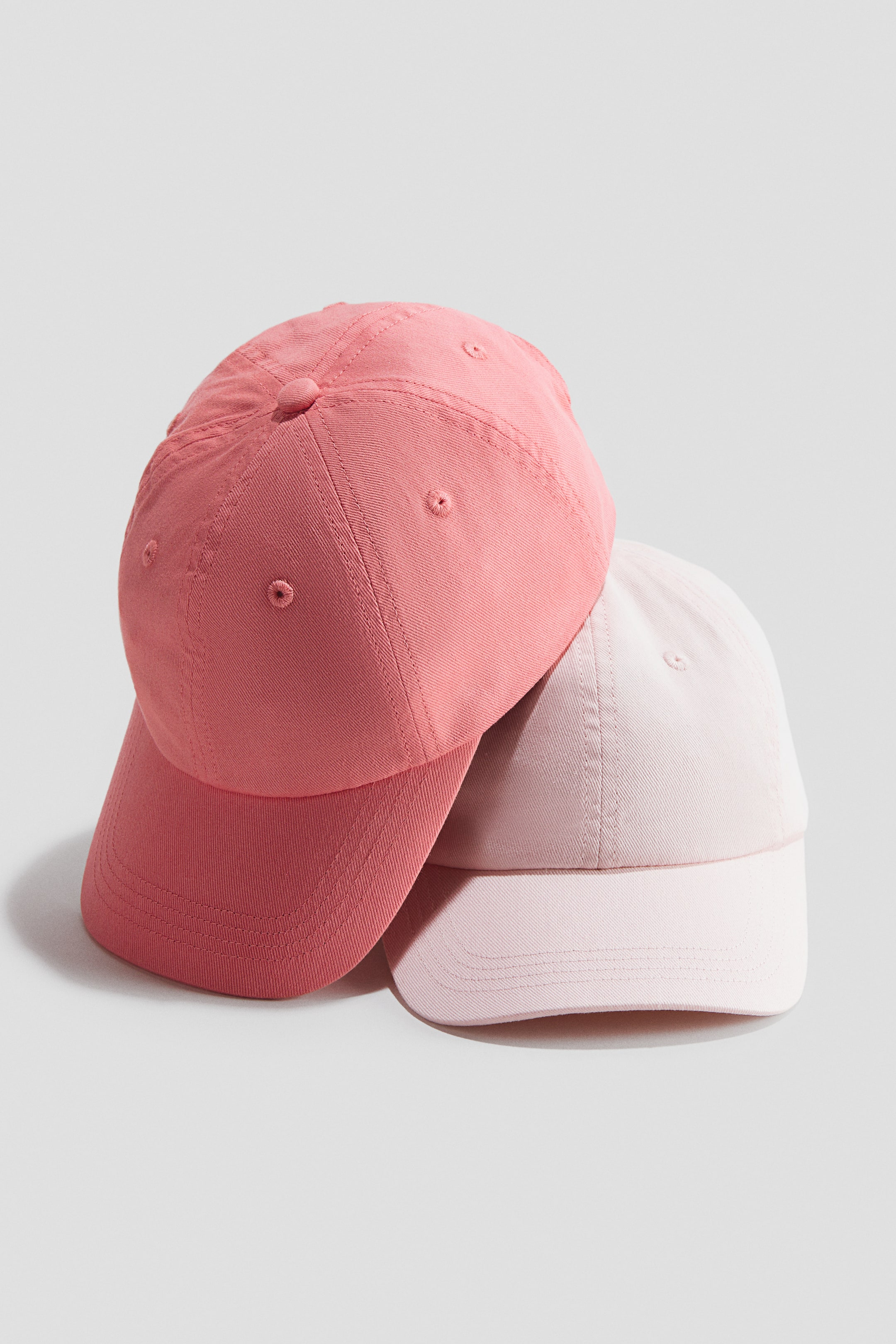 Ingrandisci l'immagine: "Due cappellini sono presentati da un'angolazione frontale, con un cappellino rosa corallo appoggiato su un cappellino rosa più chiaro. Entrambi i cappellini sono realizzati in twill di cotone, con occhielli ricamati sulle loro cupole e visiere curve."