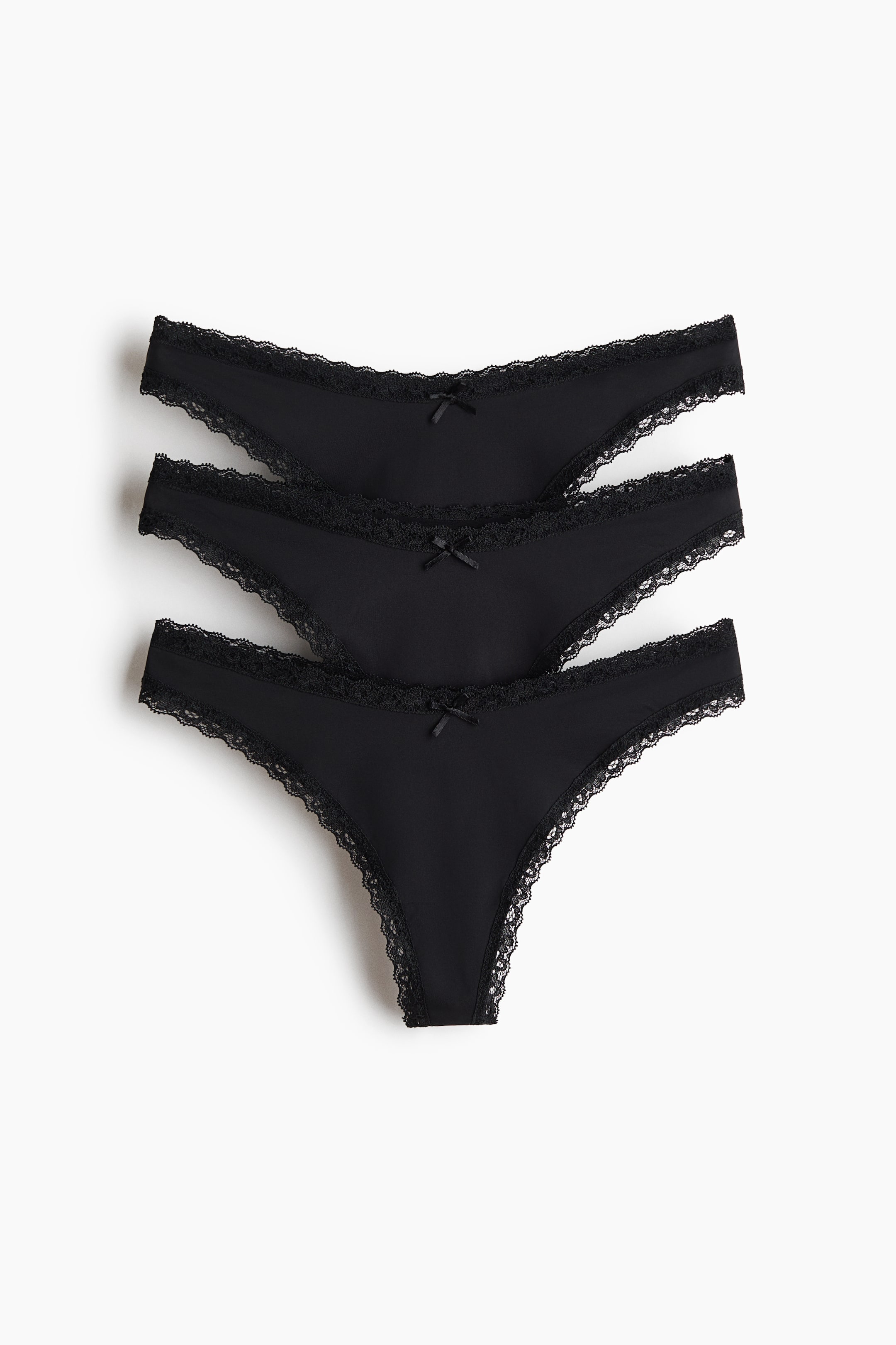 Größeres Bild anzeigen: Drei schwarze Tanga-Slips liegen in einem leicht überlappenden Stapel, schräg nach unten rechts ausgerichtet. Jeder Slip zeichnet sich durch einen tief sitzenden Schnitt, schwarze Spitzenbesätze entlang der Taille und Beinöffnungen sowie eine kleine schwarze Schleife an der Vorderseite aus.