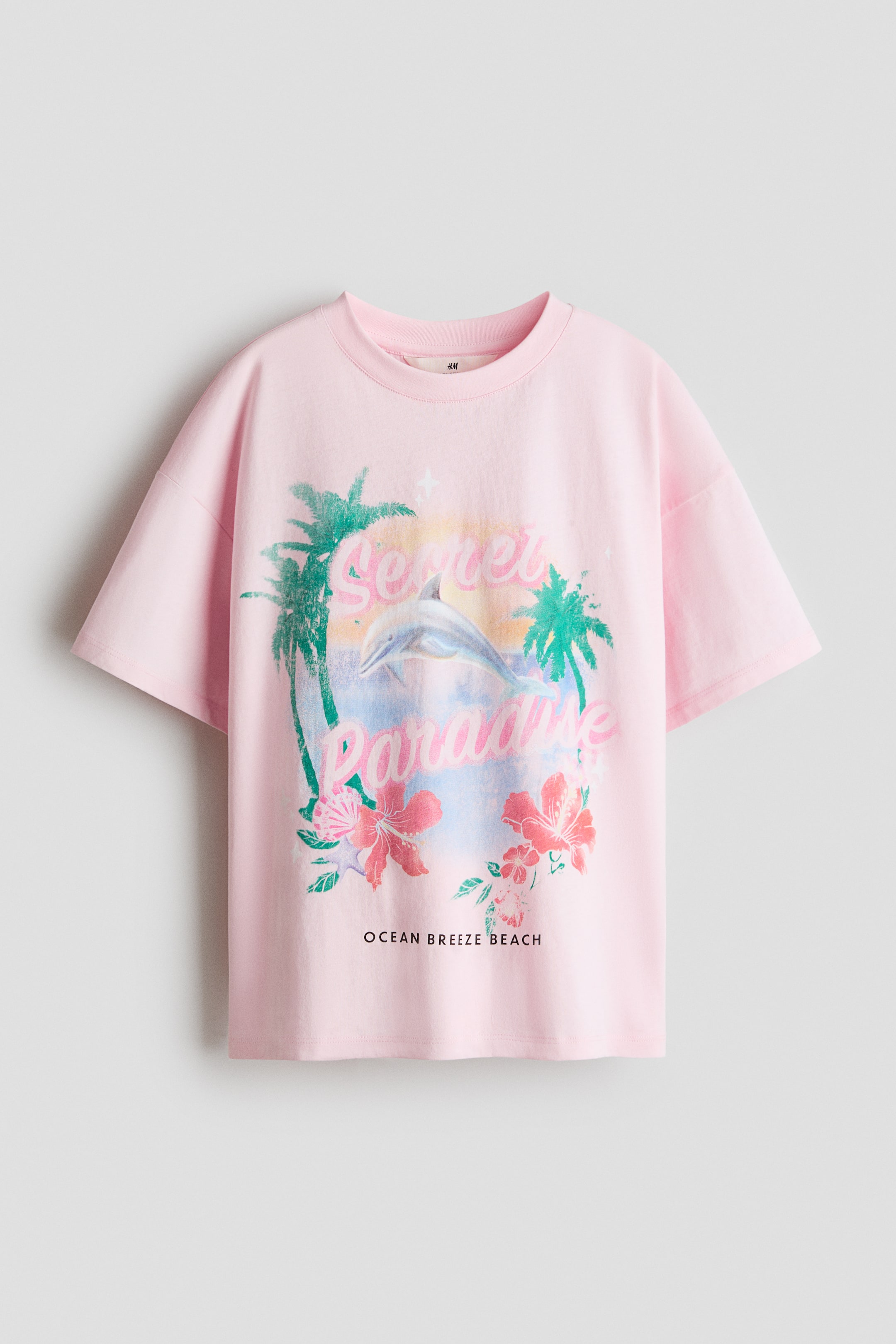 Visa större bild: En oversized rosa t-shirt i tryckt bomullsjersey visas framifrån, med en rund halsringning med ribbade kanter, nedhasade axlar och en rak fåll. Framsidan visar ett grafiskt tryck med texten "Secret Paradise", en delfin, palmer, hibiskusblommor och texten "OCEAN BREEZE BEACH".