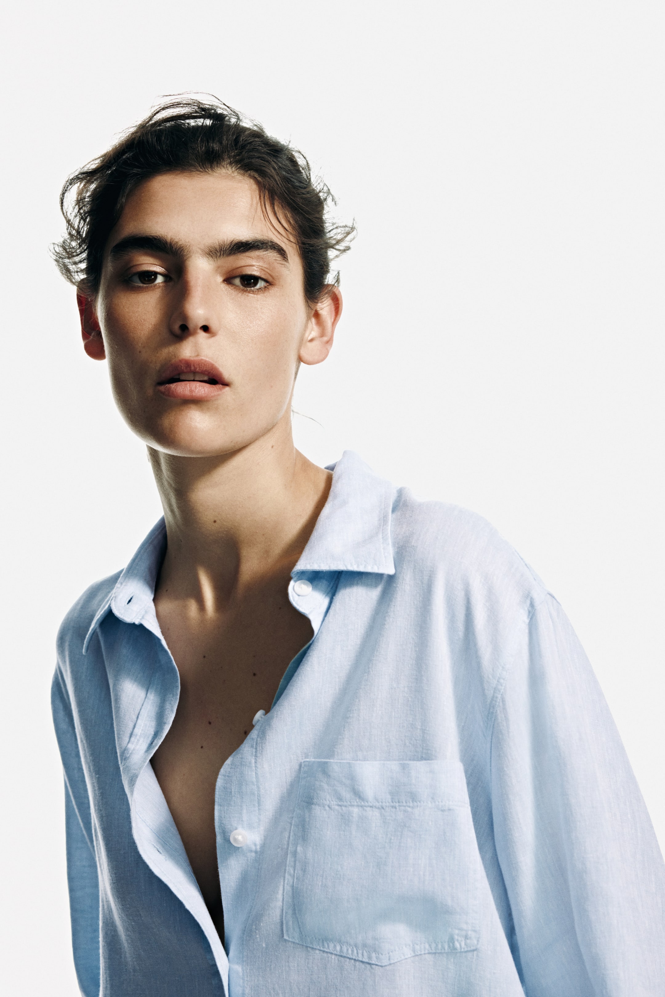 View larger image: A light blue button-up shirt, crafted from a subtly textured fabric, is showcased from a front-facing perspective. It features a pointed collar, white buttons, and a patch pocket on the left chest. The shirt is partially unbuttoned, forming a V-neck opening and offering a relaxed fit.