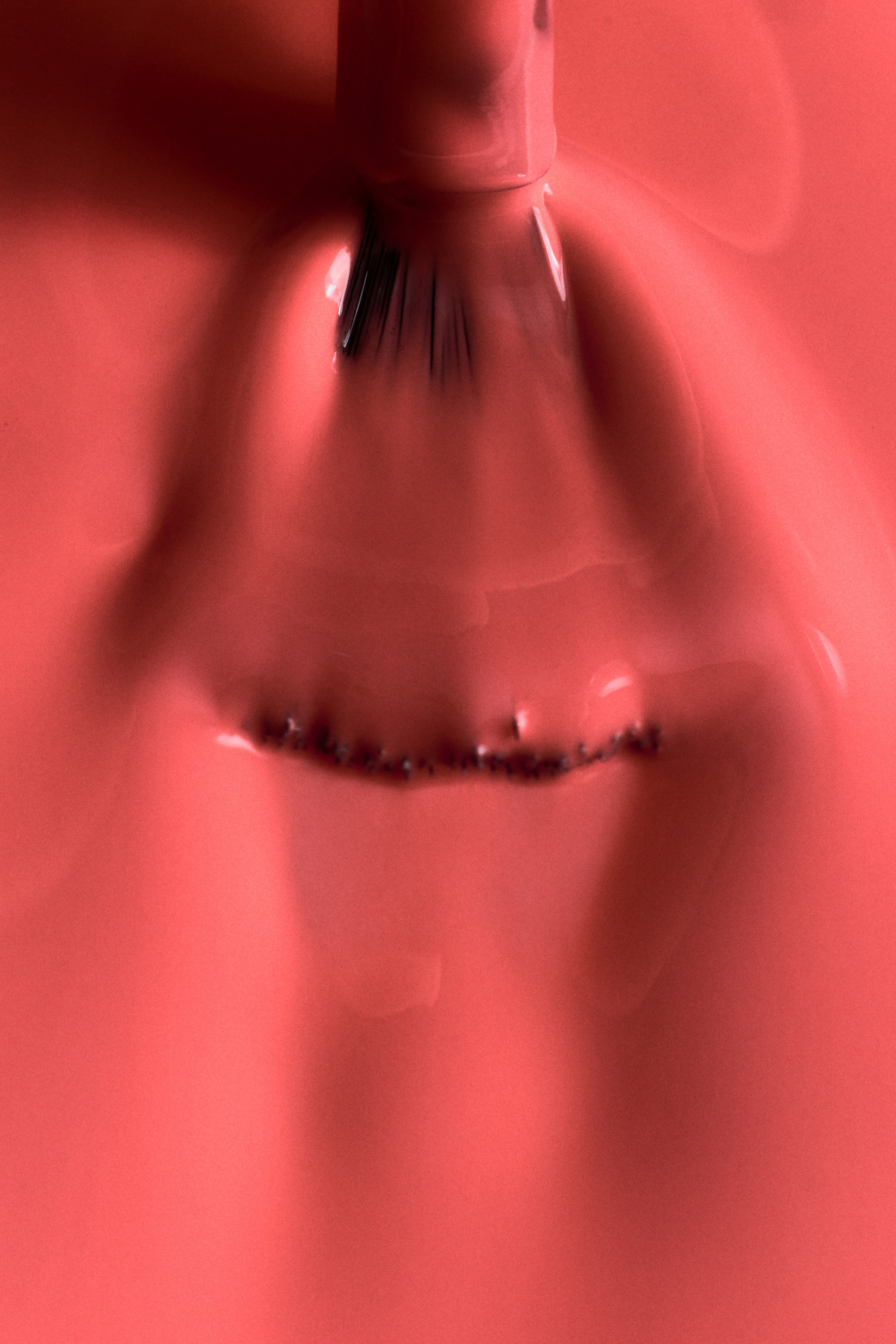 View larger image: An applicator brush is dipping into a coral-coloured nail varnish. Its fanned black bristles are submerging, creating ripples on the liquid's surface and a textured line of small dark particles just below.