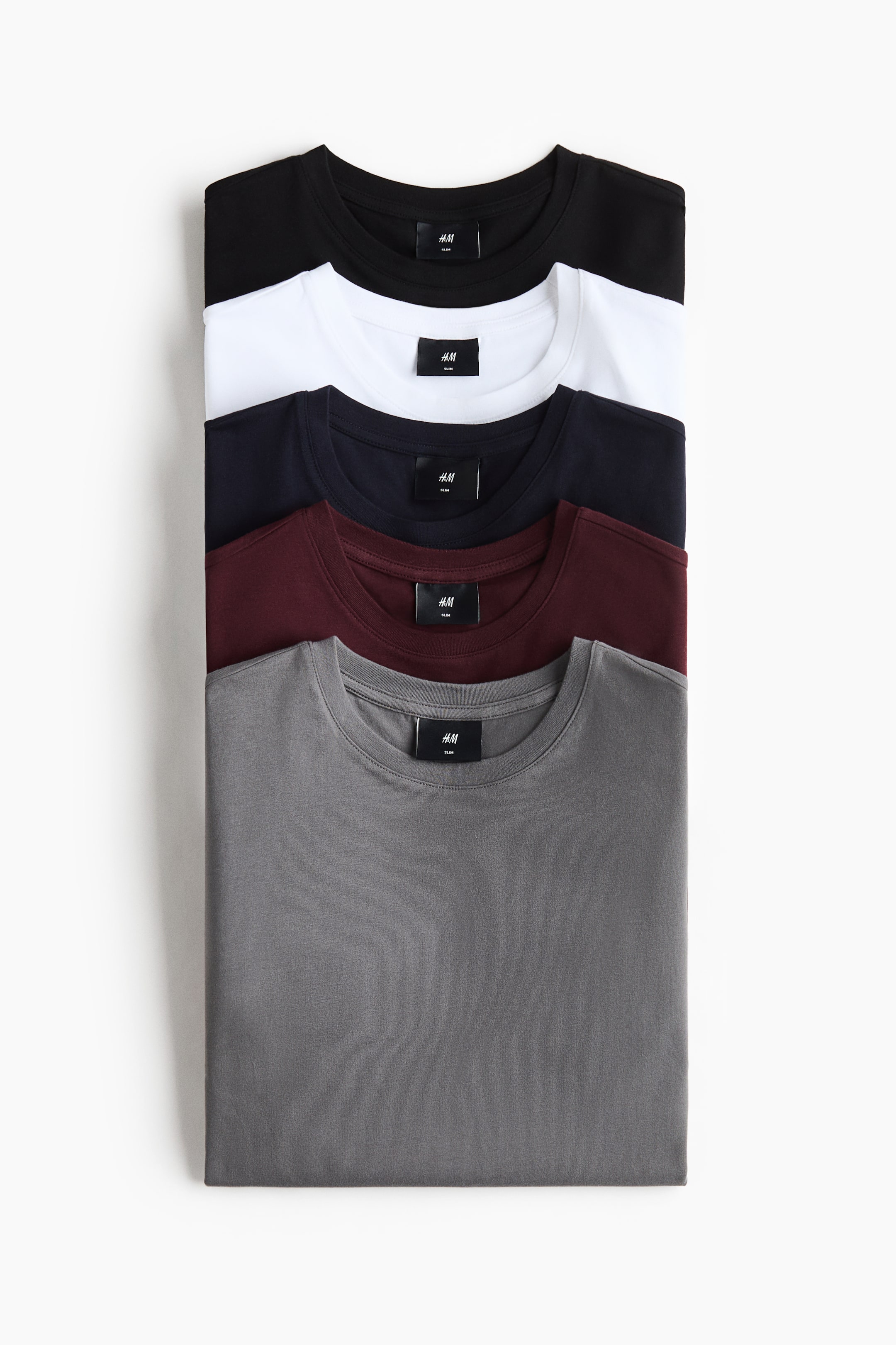 View larger image: Five T-shirts in black, white, navy, burgundy, and grey are neatly stacked, each featuring a round, rib-trimmed neckline and a black brand label.