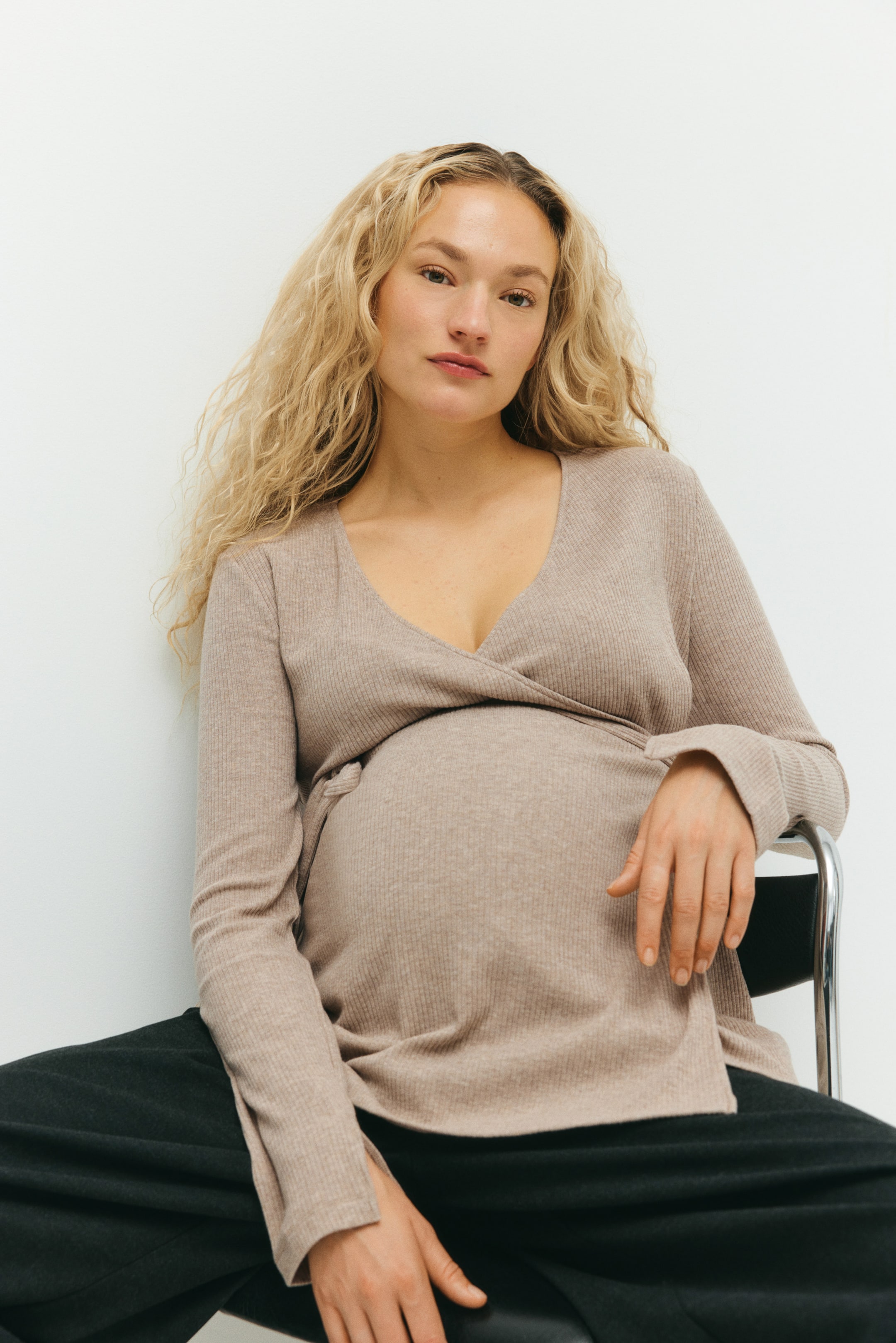 Grotere afbeelding bekijken: Een wikkeltop in zachte, geribde jersey met lange banden onder de buste en lange mouwen met een split aan de manchetten wordt gedragen door het model. Het model zit terwijl ze een donkere broek draagt, met één hand op de top rustend.