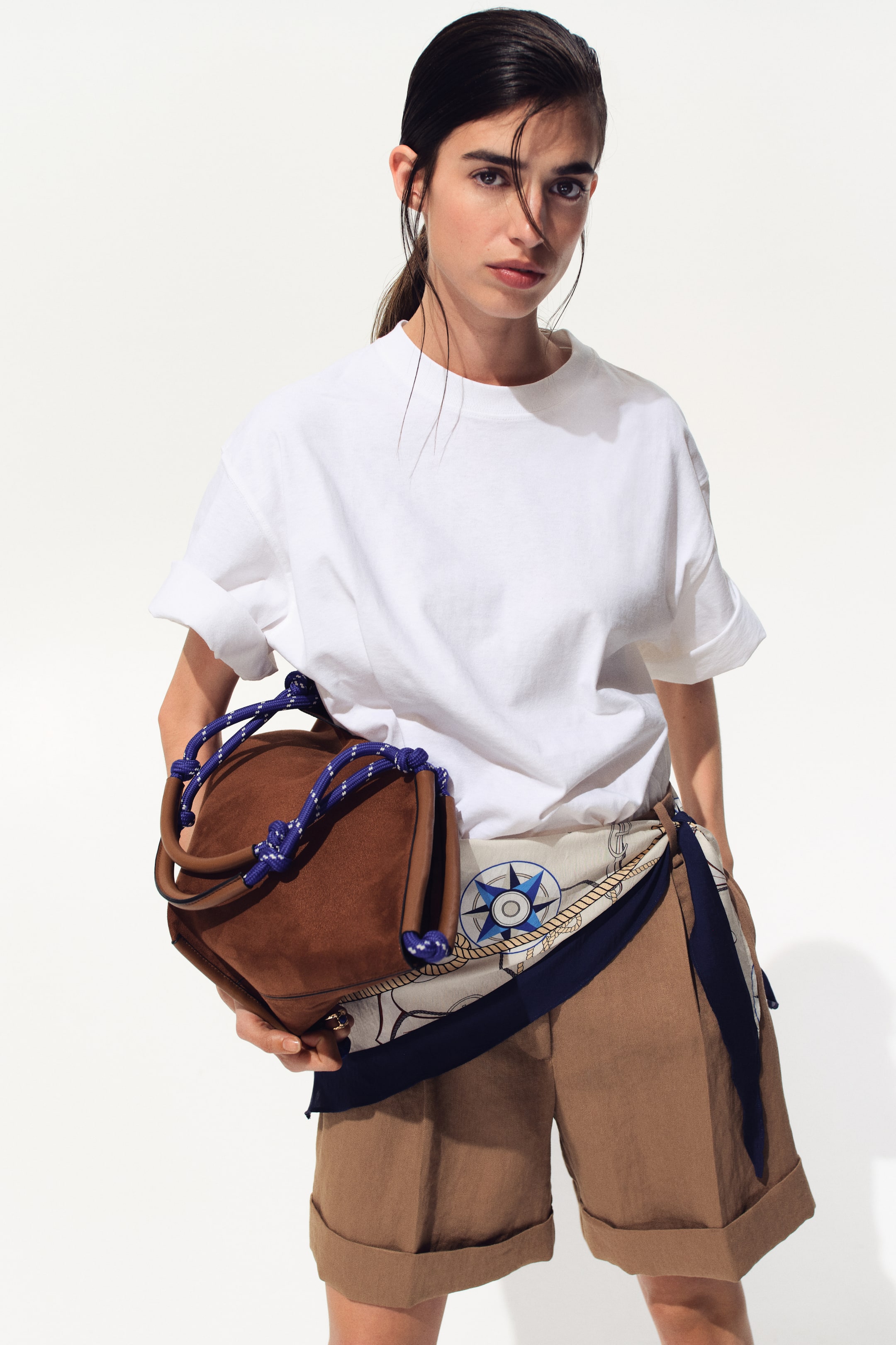 הצגת תמונה גדולה יותר: תיק יד מעור זמש חום מוחזק, עם חבל קלוע בכחול ולבן המתפתל בצורה מורכבת סביב שתי ידיותיו החומות המעוגלות ויוצר קשרים דקורטיביים בחלק העליון.