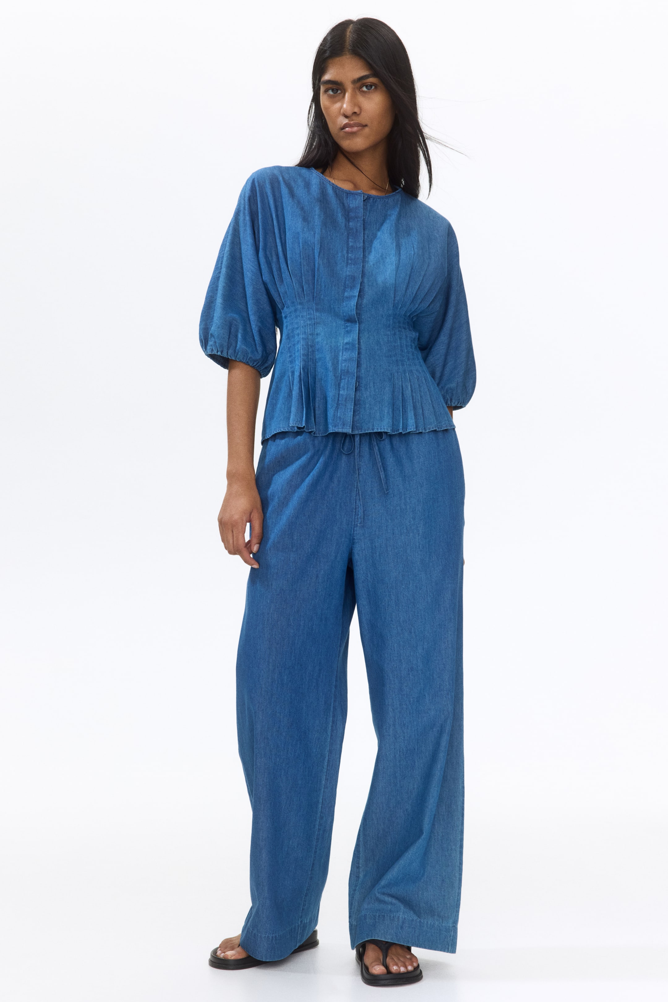 View larger image: A blue chambray-like top features a round neck, a button-front placket, and voluminous sleeves, showcasing a fitted bodice with vertical pleats leading to a pleated peplum hem. The matching wide-leg pants feature a drawstring elasticated waist.