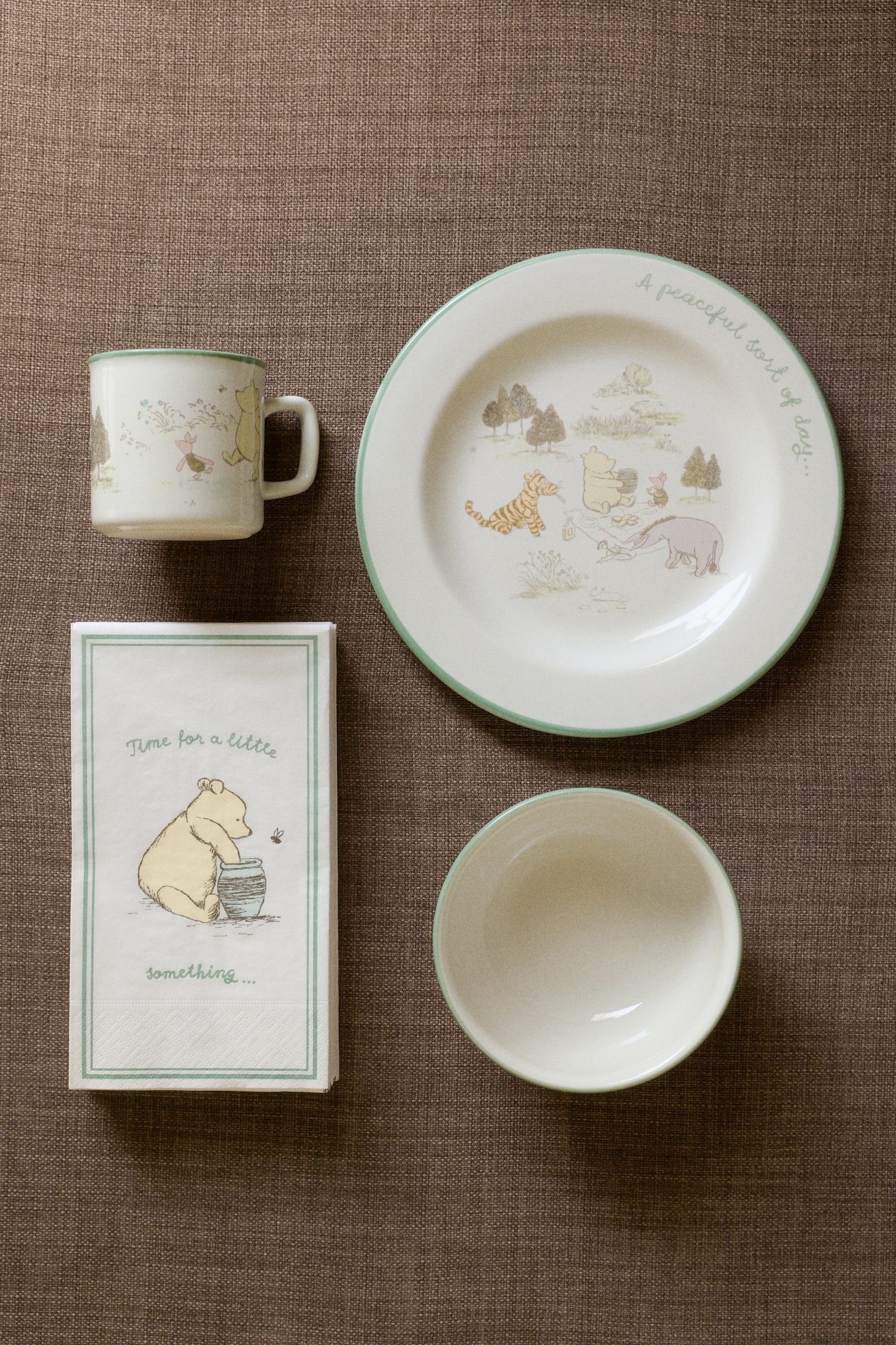 A Winnie the Pooh dining set featuring a white mug with a green rim and character illustration, a white plate with a green rim and a detailed picnic scene, a stack of white napkins with a green border and a Pooh with a honey pot illustration, and a white bowl with a green rim. - 1