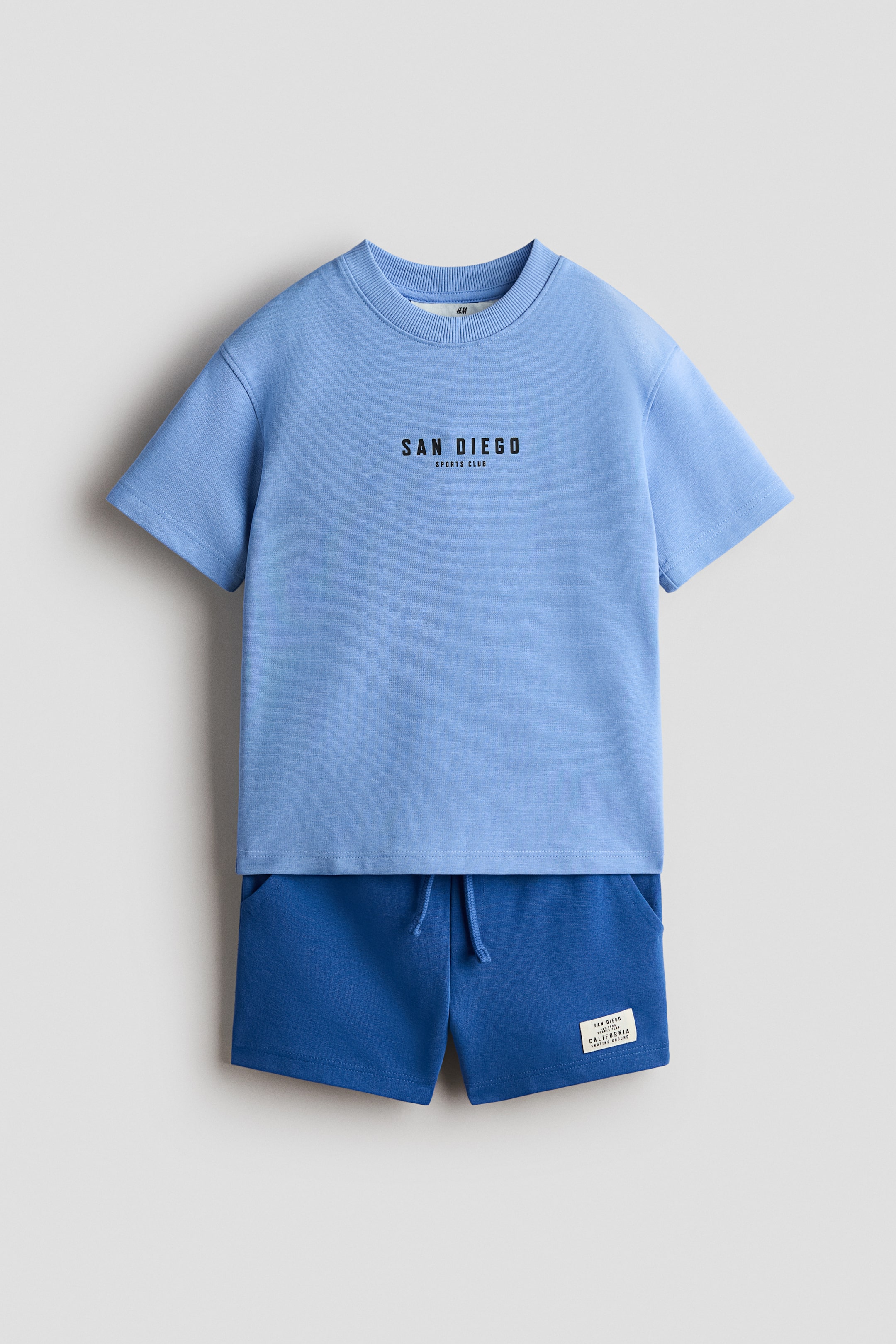 View larger image: A light blue t-shirt and darker blue shorts set is lying flat, presented from the front. The t-shirt features a ribbed neckline, gently dropped shoulders, and "SAN DIEGO SPORTS CLUB" text across the chest, while the shorts display an elasticated, drawstring waist, a fake fly, diagonal side pockets, and a small rectangular label on the right leg.