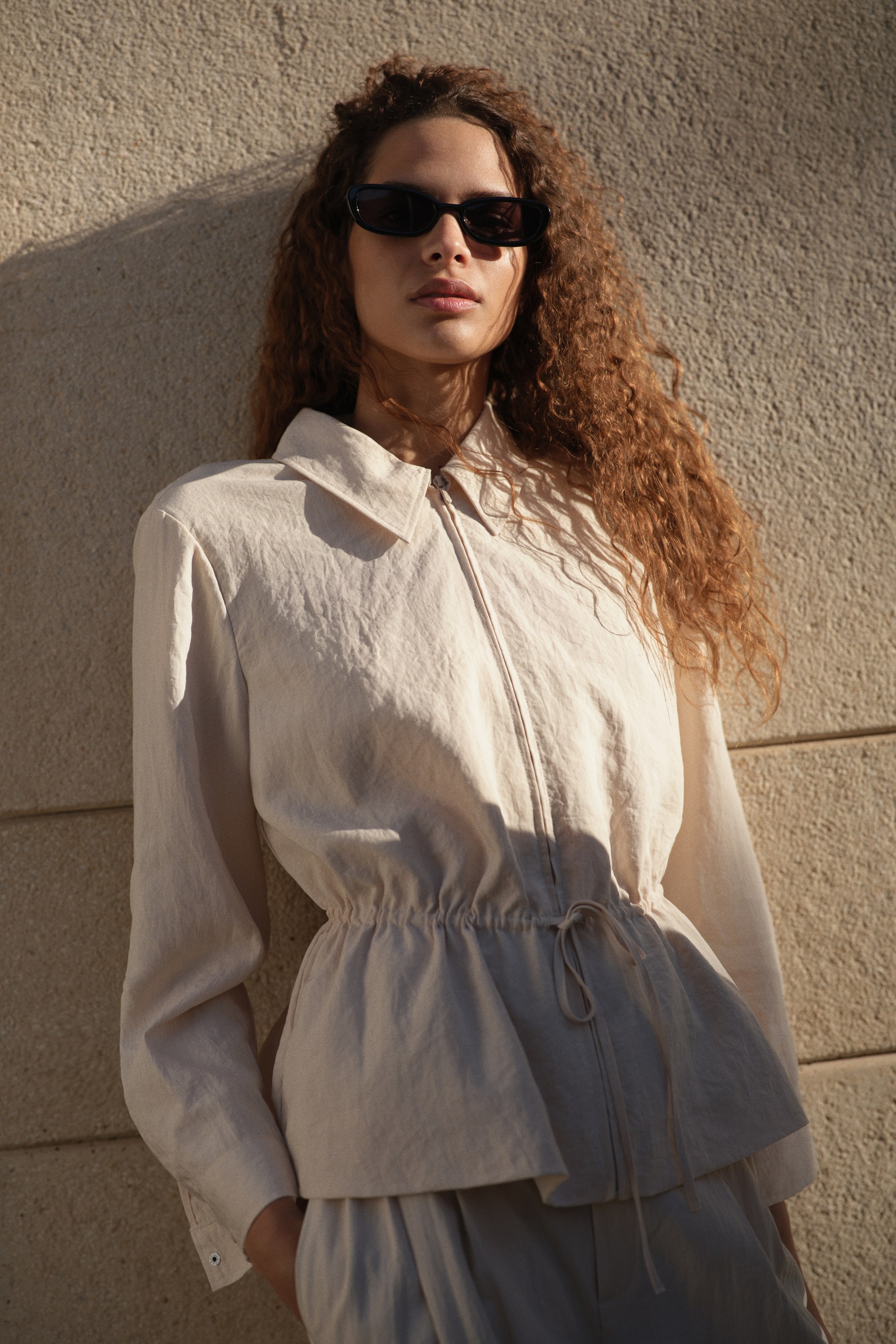 View larger image: A light beige long-sleeved top, crafted from a textured fabric, features a pointed collar, a full-zip front, and buttoned cuffs, presented from a slight angle. Its drawstring waist cinches to create a peplum hem, and it is paired with matching loose-fitting trousers.