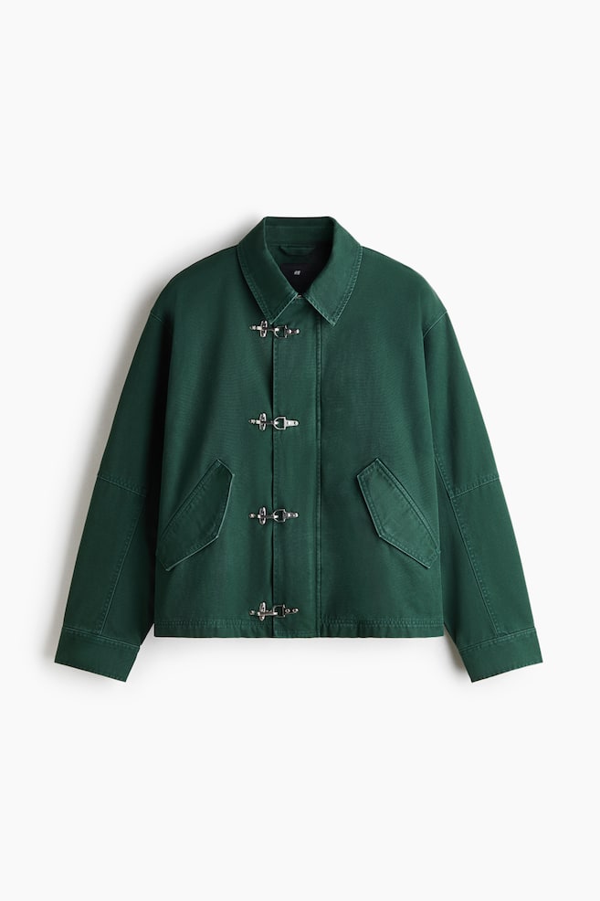 A dark green cotton canvas jacket is facing front, showcasing a classic point collar, dropped shoulders, and a front wind flap secured with four silver metal clasps. The jacket features two angled, jetted front pockets with pointed flaps. - 1