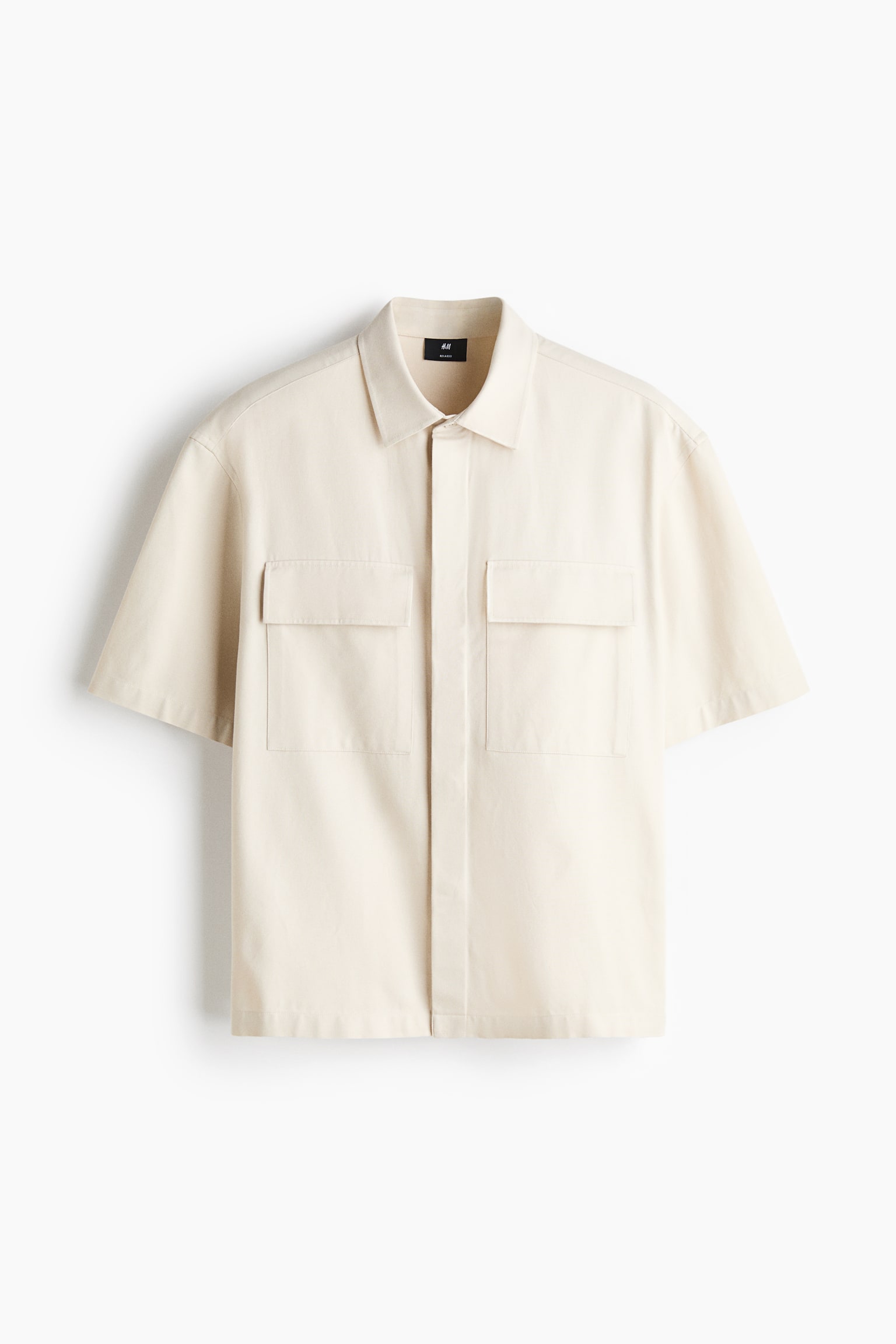 A cream-colored short-sleeve shirt is facing front, displaying a classic collar, a concealed button placket, and two large patch chest pockets with flaps. The shirt is showcasing a relaxed fit with a straight-cut hem.
