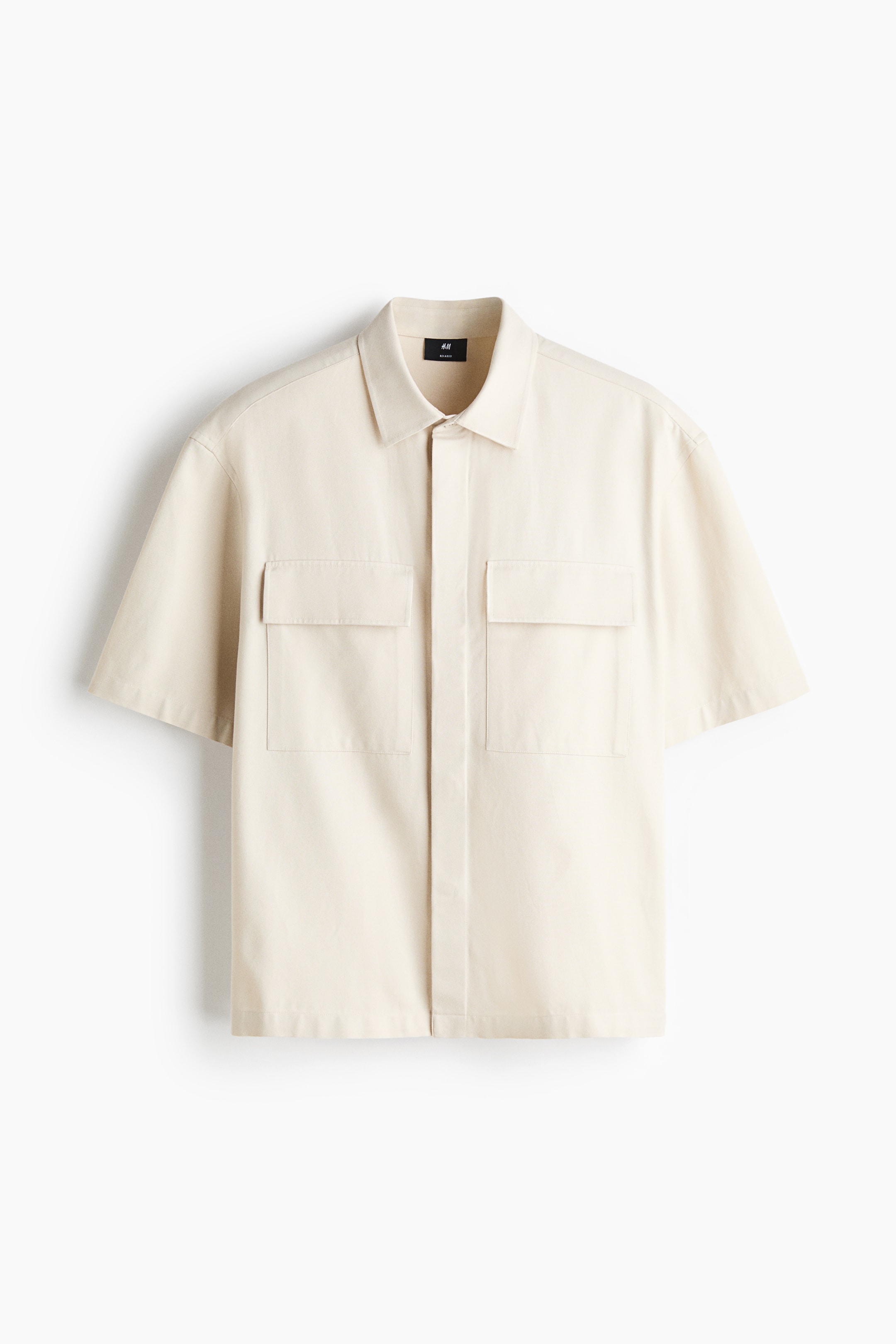 View larger image: A cream-colored short-sleeve shirt is shown from the front, featuring a classic collar, concealed button placket, and two large flap patch chest pockets. The shirt has a relaxed fit with a straight-cut hem.