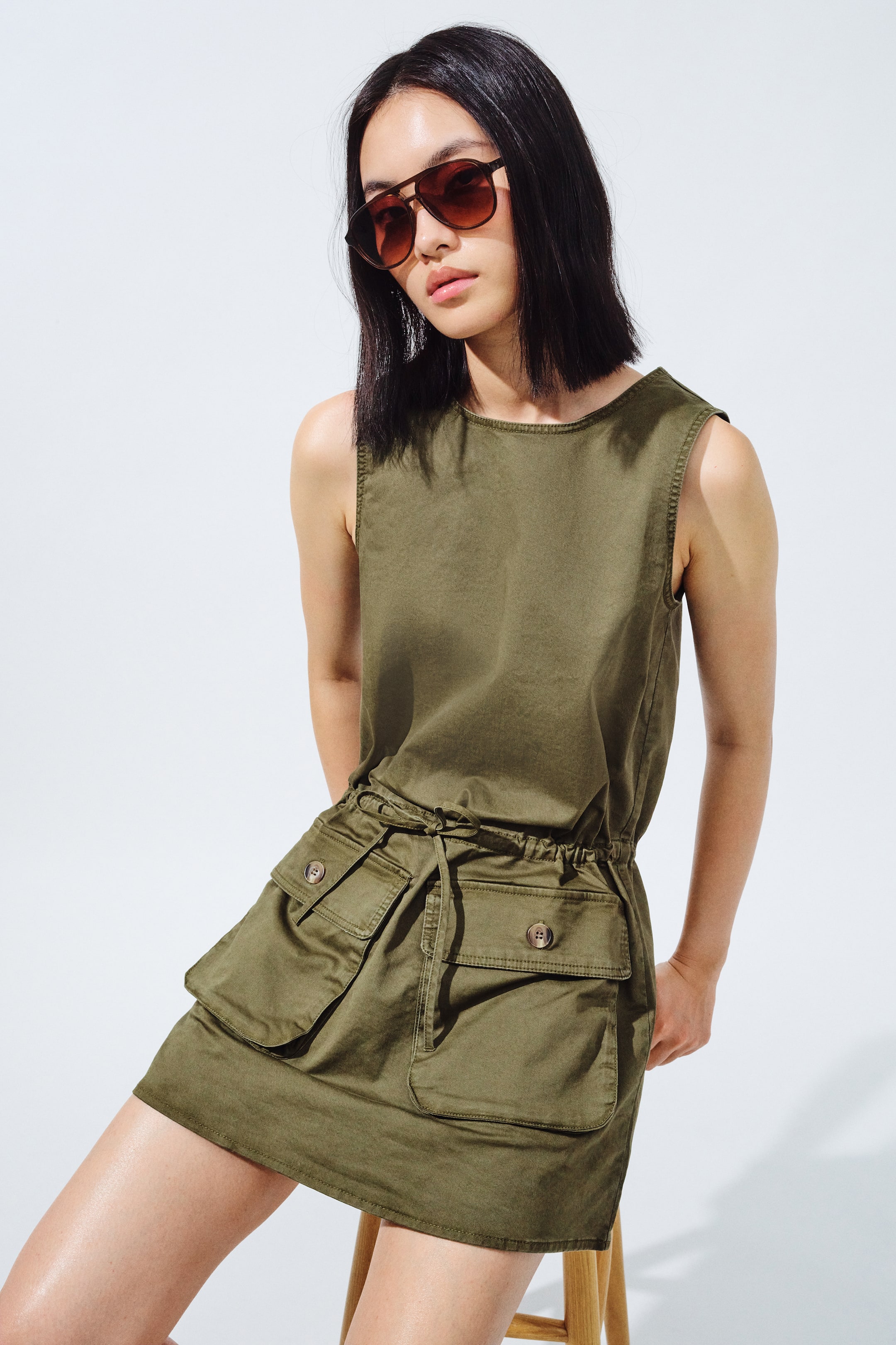 View larger image: This olive green sleeveless utility dress features a crew neck and a drawstring waist that cinches the silhouette. The front skirt is detailed with large flap pockets and visible buttons, ending just above the knee.