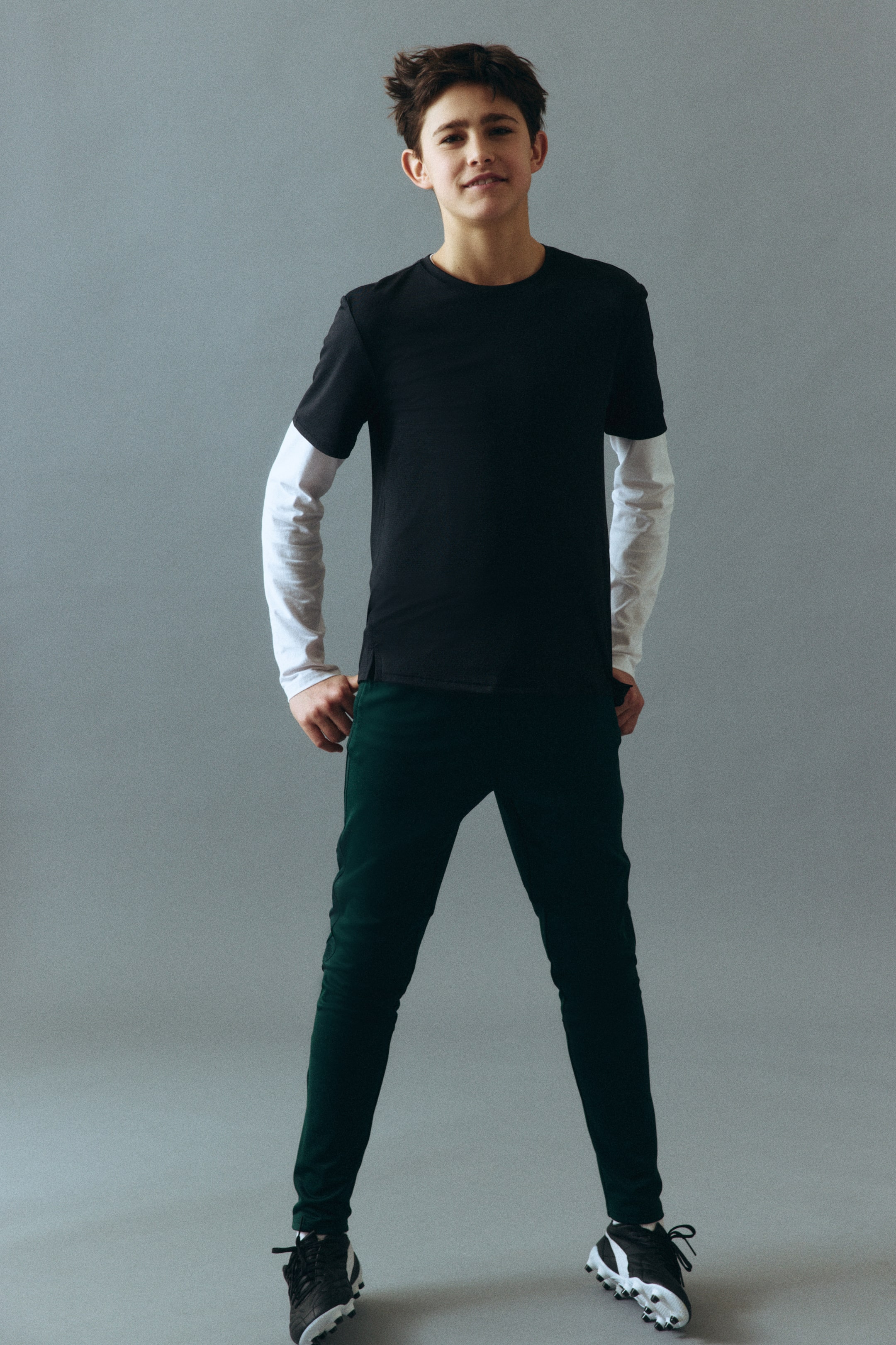 Visa större bild: Outfiten, sedd snett framifrån, består av en svart crewneck-t-shirt som bärs över en vit långärmad tröja. Mörkgröna slim-fit-byxor kompletterar helheten, och svarta och vita sportskor med dubbar på sulorna bärs också.