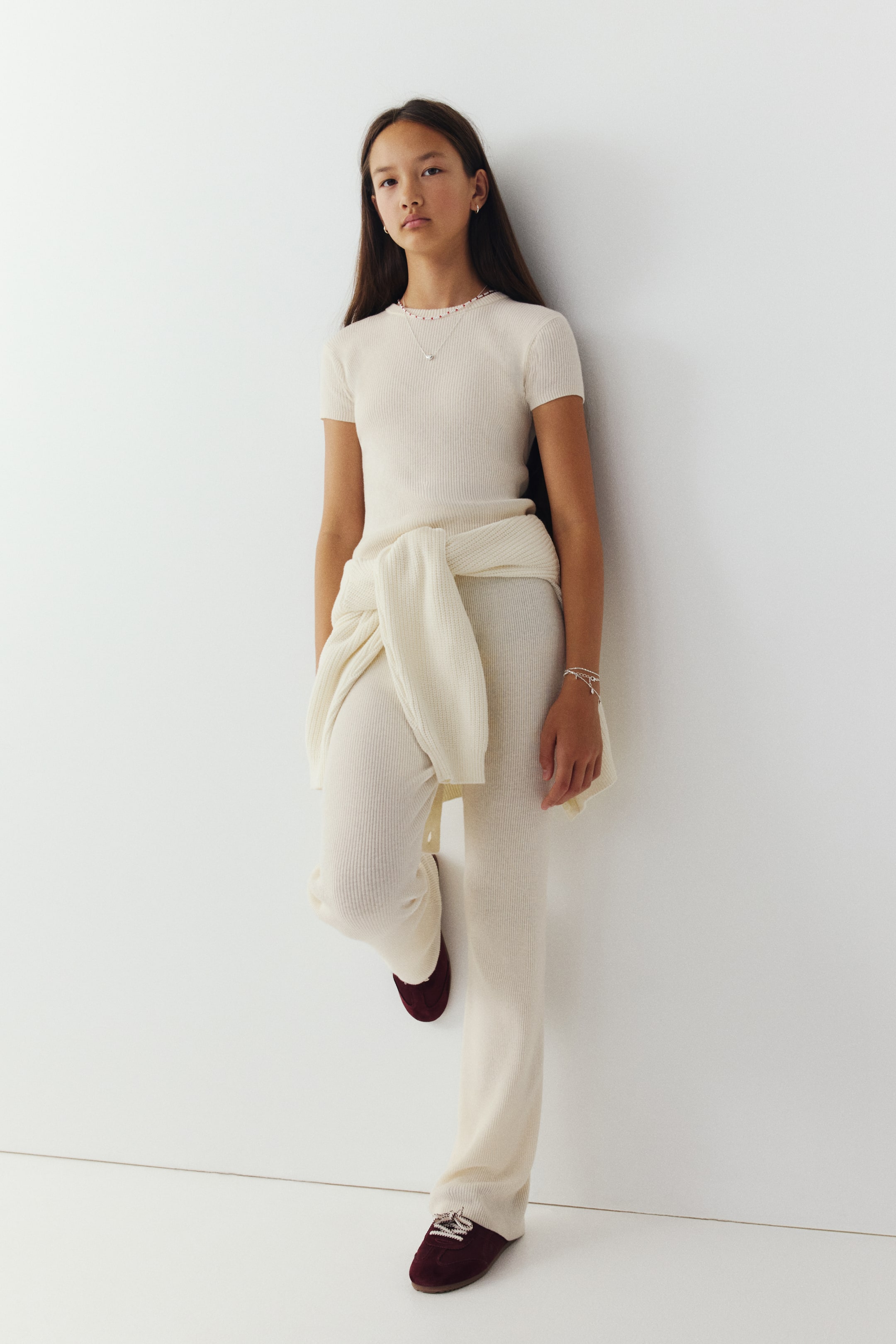 View larger image: A cream-coloured rib-knit set is shown from the front, featuring a short-sleeved, round-neck top and matching wide-leg trousers. A coordinating knitted sweater is tied around the waist of the trousers, which are styled with dark red trainers.