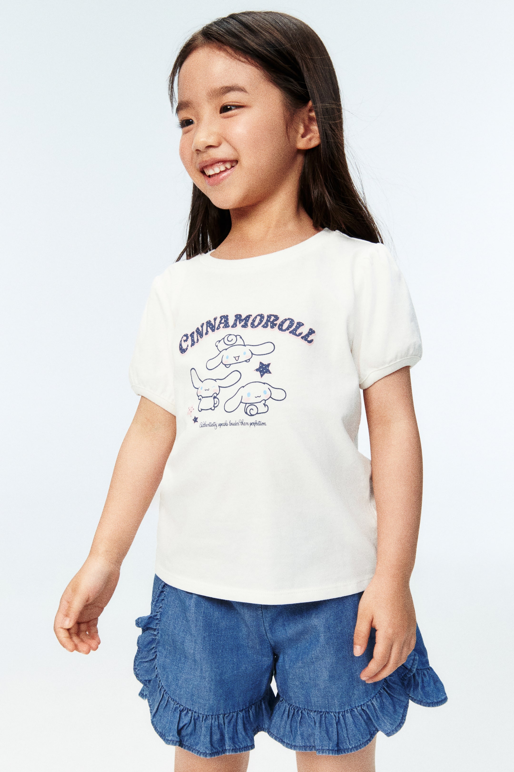 Xem ảnh lớn hơn: Người mẫu đang mặc áo thun trắng có họa tiết in Cinnamoroll màu xanh ở mặt trước. Áo có cổ tròn viền gân và tay ngắn phồng với bo tay hẹp, dệt gân. Người mẫu cũng đang mặc quần short xanh có bèo nhún.