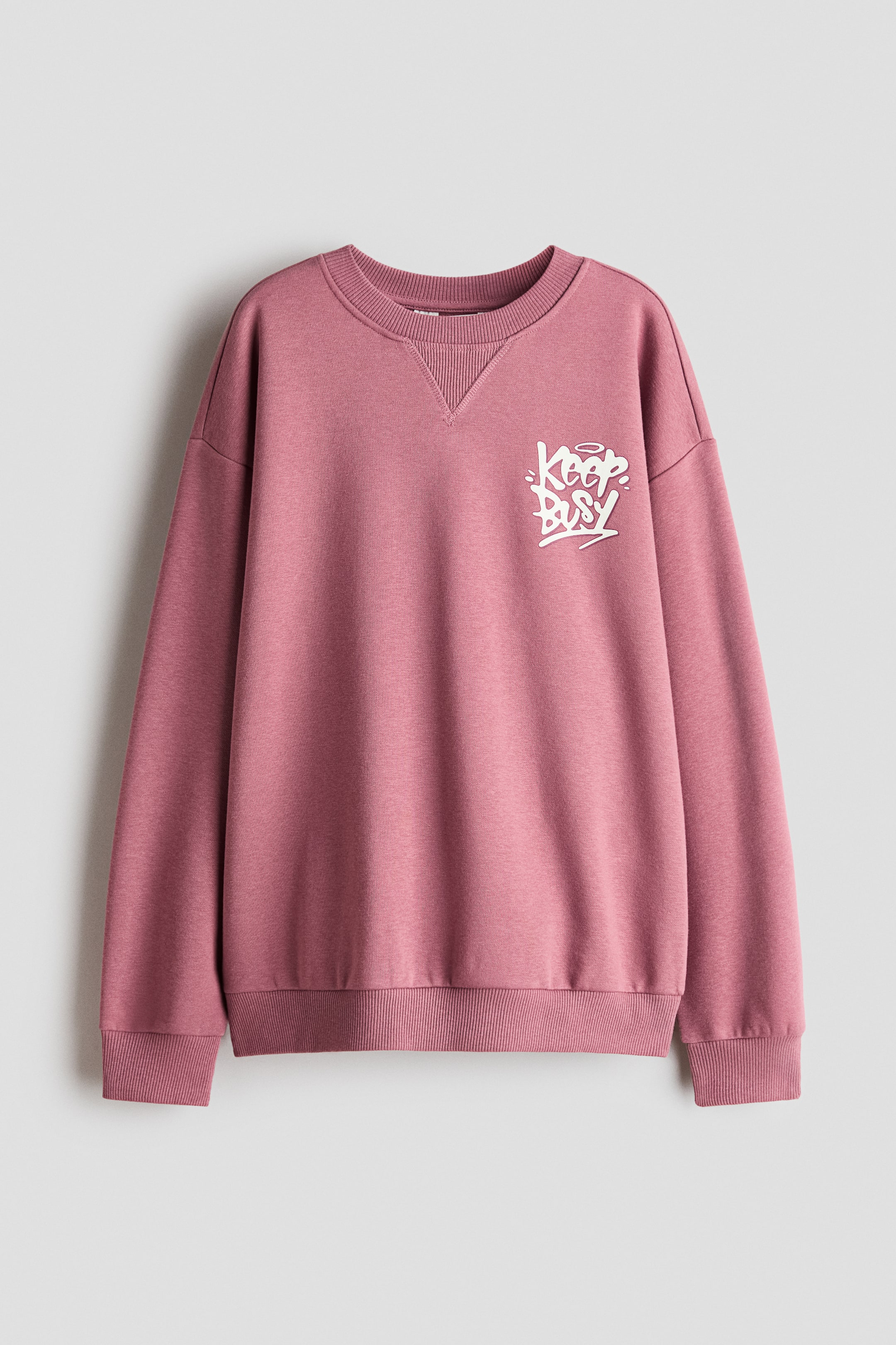 Agrandir l'image: Ce sweat-shirt rose poudré est présenté de face, avec un col rond côtelé orné d’un détail en V, des épaules tombantes, ainsi que des poignets et un ourlet côtelés. Un petit motif graphique blanc de style graffiti, portant l’inscription « Keep Busy », décore la poitrine gauche.