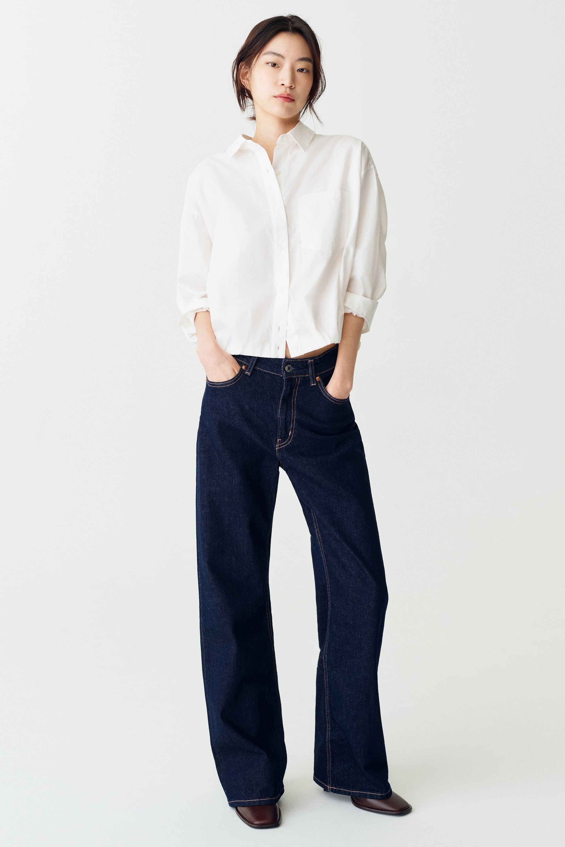 Xem ảnh lớn hơn: Một chiếc áo sơ mi cotton trắng có cổ áo, hàng cúc cài phía trước, một túi ngực và gấu áo phồng kiểu bubble đang được người mẫu mặc. Áo được phối với quần jeans ống rộng màu xanh đậm, người mẫu đứng với hai tay đút túi.