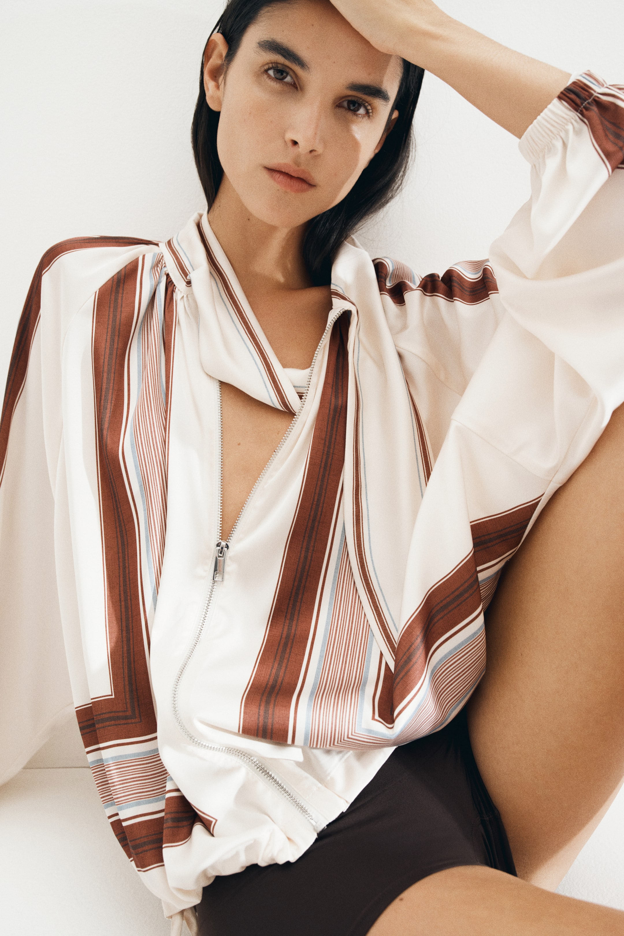Größeres Bild ansehen: Eine übergroße Jacke aus weich fallendem Creme-Satin mit braunen und blauen Streifen wird vom Model getragen. Sie verfügt über einen Schal-Kragen, einen teilweise geöffneten Reißverschluss, Raglanärmel mit elastischen Bündchen und einen gerafften Saum mit einem versteckten Kordelzug. Geraffte Nähte erzeugen einen drapierten Effekt im gesamten Design, und der erhobene Arm des Models hebt das elastische Bündchen hervor, während ein weißes Oberteil unter der geöffneten Jacke sichtbar ist.
