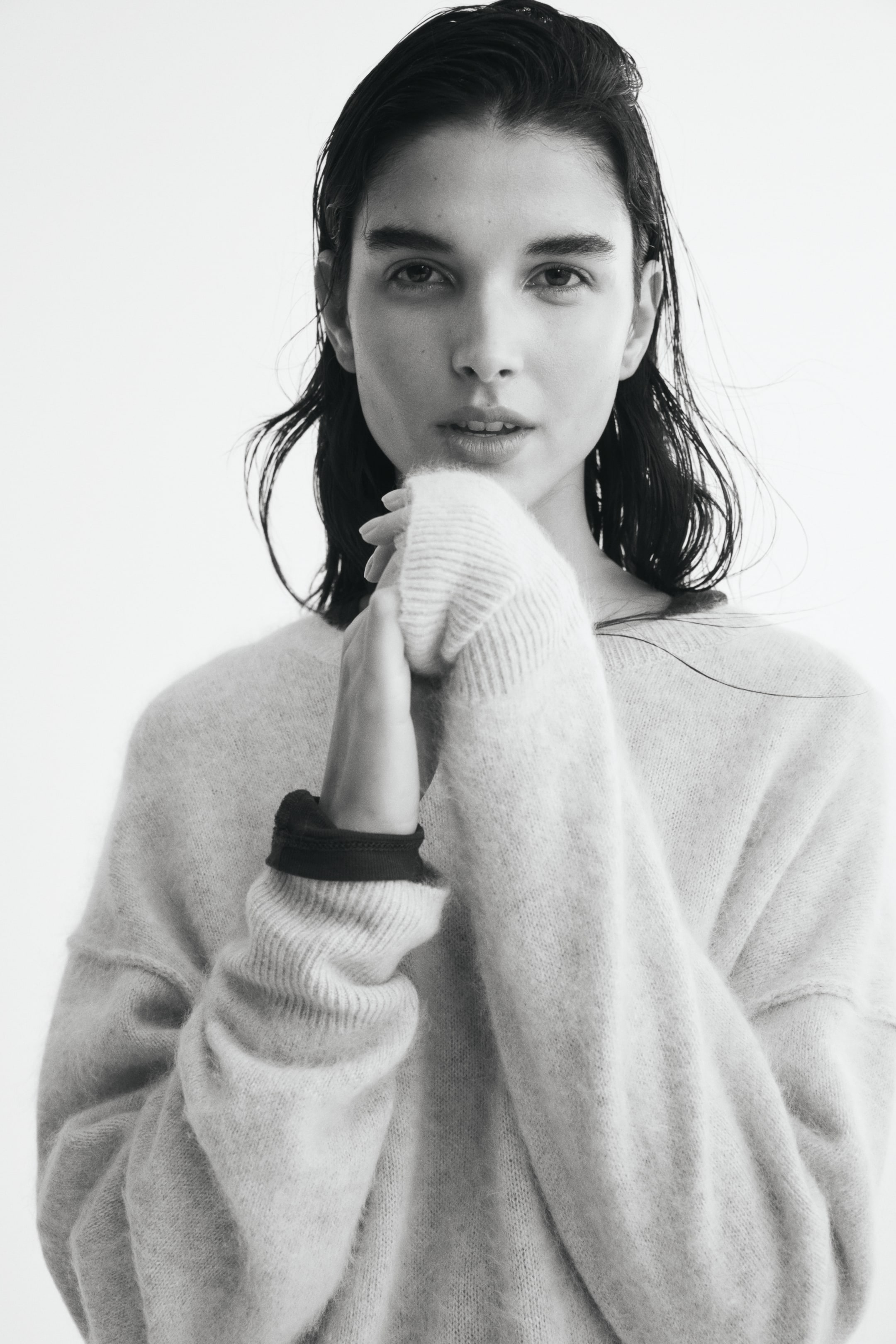 Größeres Bild ansehen: Ein weicher Strickpullover aus Wolle mit Rundhalsausschnitt, überschnittenen Schultern und langen Ärmeln mit Rippbündchen wird vom Model getragen. Das Model hält eine Hand nah am Kinn und zieht das lange Rippbündchen über die Finger, während der andere Ärmel leicht zurückgeschoben ist und am Handgelenk eine dunkle Unterschicht zum Vorschein bringt.