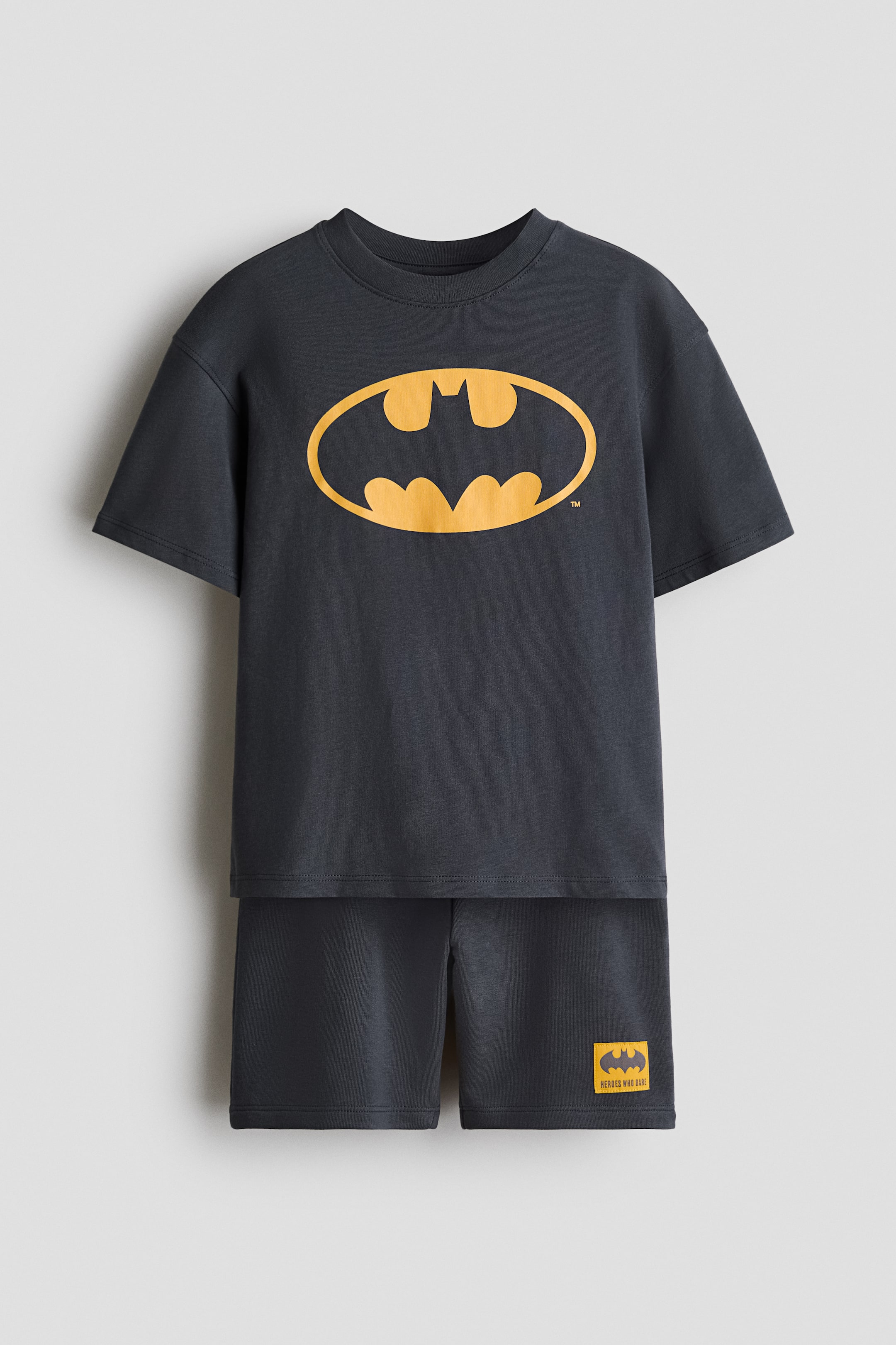 Se større billede: Et mørkegråt todelt sæt ligger fladt, med en kortærmet t-shirt, der har et stort gult Batman-logo på brystet. De matchende shorts har en lille gul mærkat med et Batman-logo og "HEROES WHO DARE!" på det nederste højre ben.