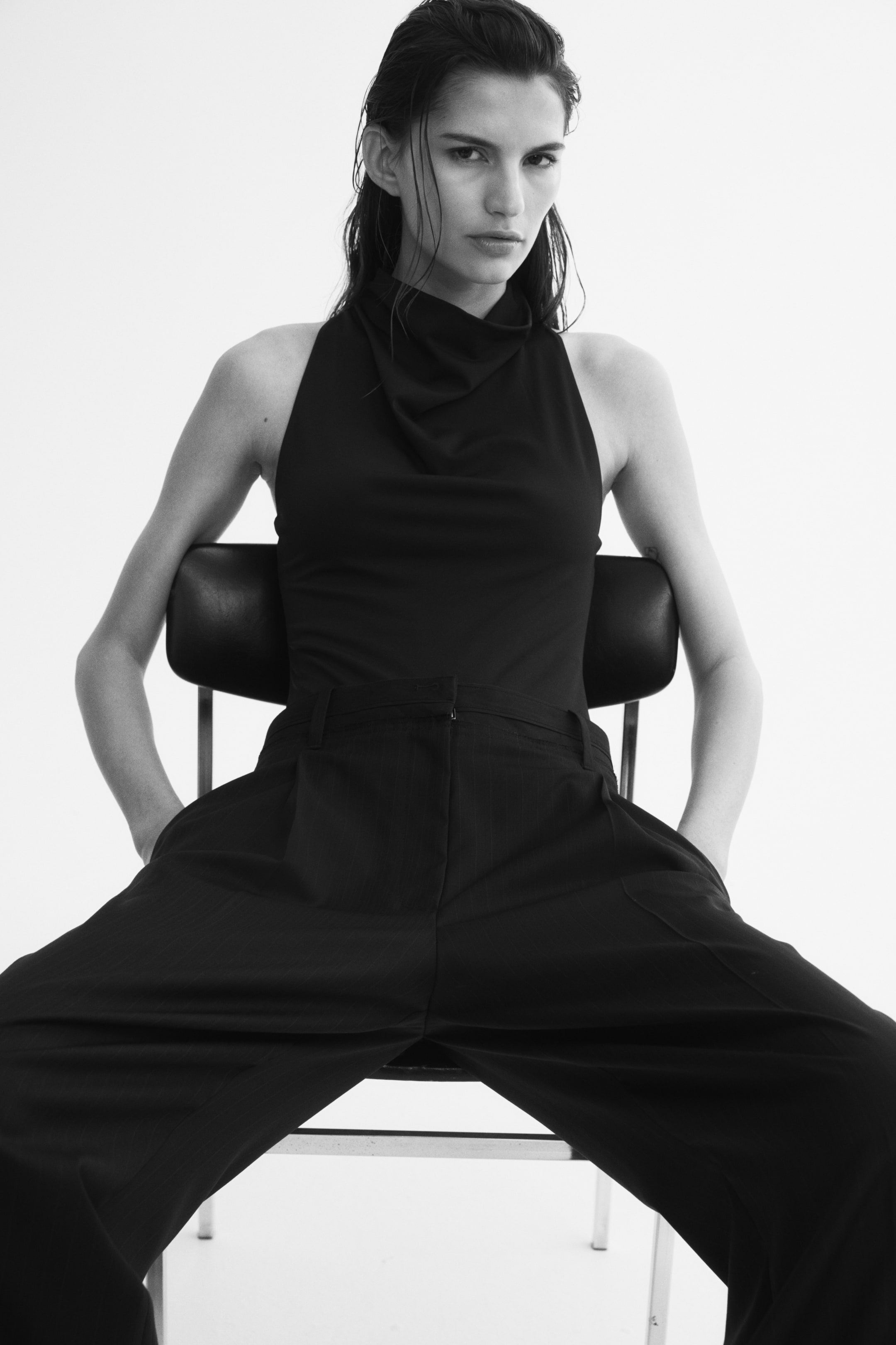 View larger image: A sleeveless, dark-coloured bodysuit featuring a draped cowl neck and a narrow cut at the shoulders is being worn by the model. The fitted bodysuit is styled tucked into a pair of wide-leg trousers.