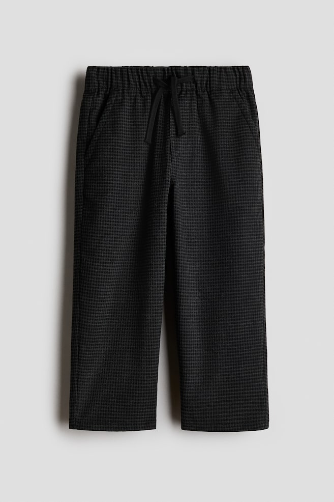 A pair of dark grey trousers featuring a fine houndstooth pattern is shown facing front, showcasing an elasticized waistband with an adjustable black drawstring. The trousers are falling into a straight-leg silhouette and include side-seam pockets. - 1